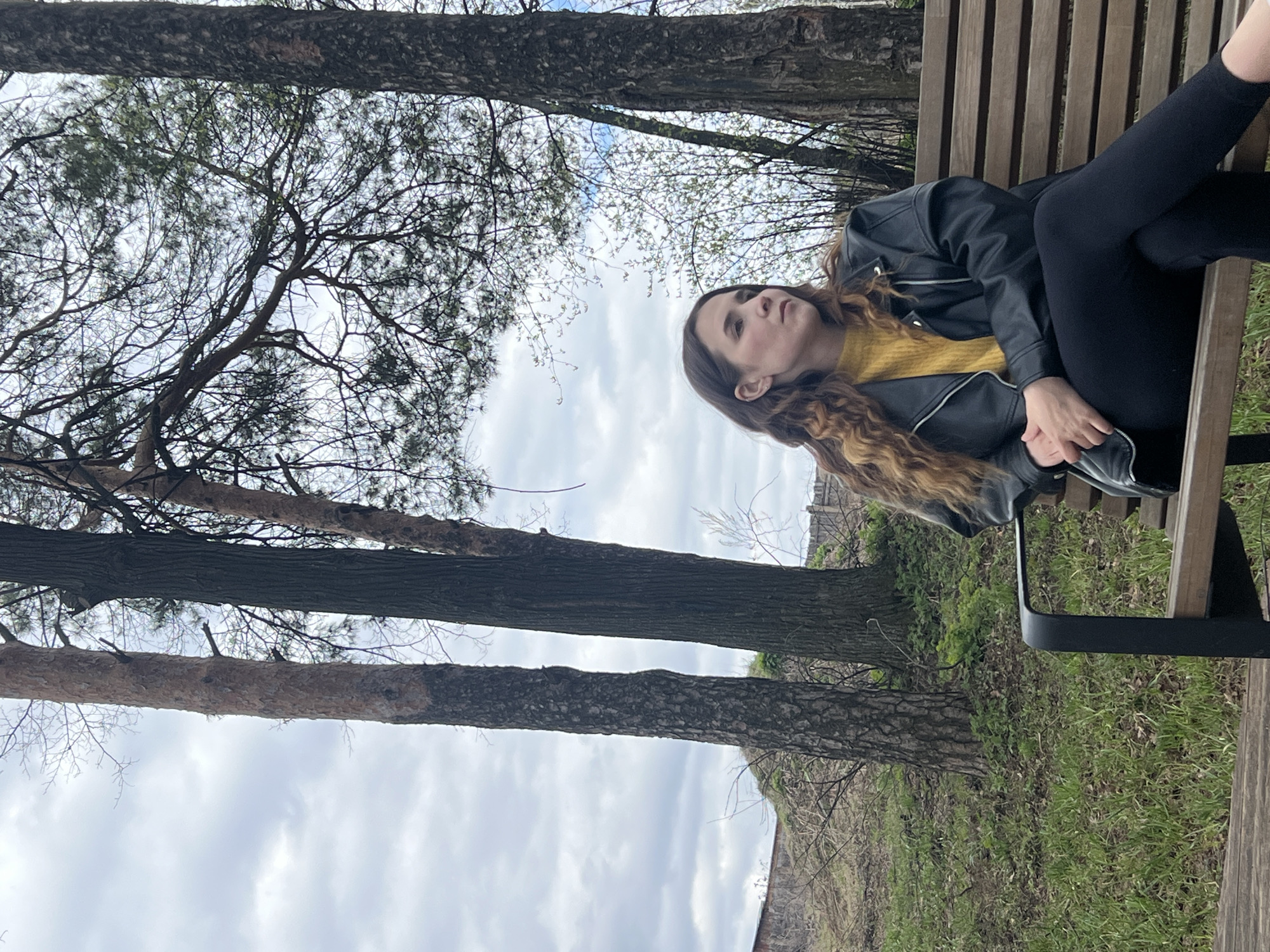 The image, taken in Votkinsk, Russia, captures a young woman reclining on a wooden park bench in an outdoor, natural setting. The woman is positioned on the right side of the frame, lying horizontally with her legs crossed. She has long, wavy brown hair, and is dressed in a black faux-leather jacket over a mustard yellow top, paired with black leggings. Her gaze is directed upwards and slightly to her right, conveying a calm or contemplative mood.
The scene is dominated by several tall, sturdy trees with rough bark on the left side and in the background. Their branches, some appearing bare or with nascent spring foliage, stretch across an overcast sky filled with clouds. The ground around the bench is covered with green grass and some scattered dry leaves, suggesting late spring or early autumn. The overall impression is a peaceful daytime scene under a bright, cloudy sky. No text is visible in the image.