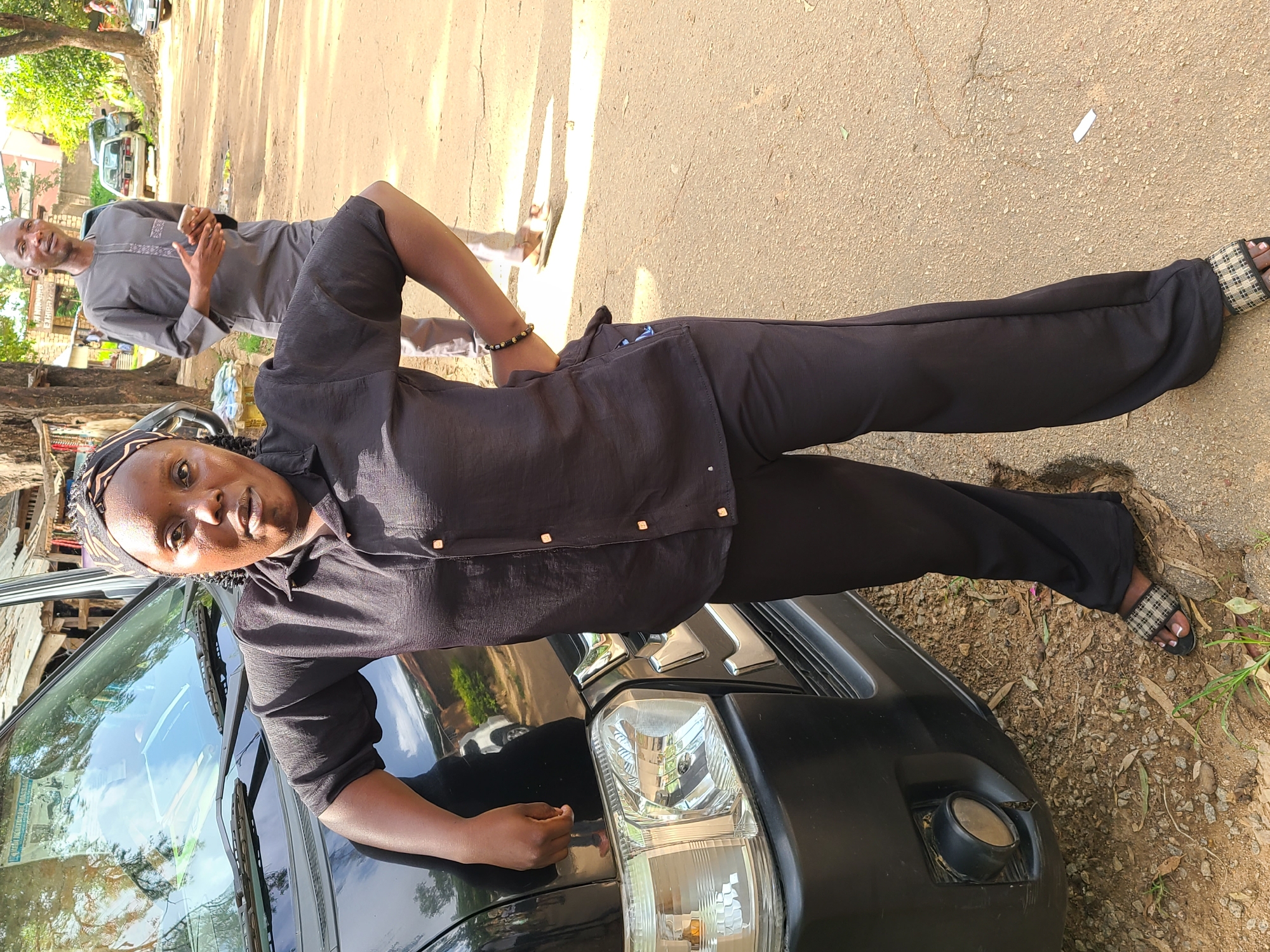 A dark-skinned woman stands next to a black van, her left arm resting on the hood. She is wearing a black collared shirt with gold buttons and matching black pants. A patterned headscarf covers her hair, and she has a bracelet on her right wrist. Her feet are clad in black and white checkered slides.
In the background, a man in a grey tunic and trousers holds a mobile phone, looking to his left. The scene appears to be an outdoor setting, possibly a roadside in Jos, Nigeria, with dirt ground and some vegetation visible. Other vehicles and structures can be seen in the distance. The lighting suggests it is daytime.