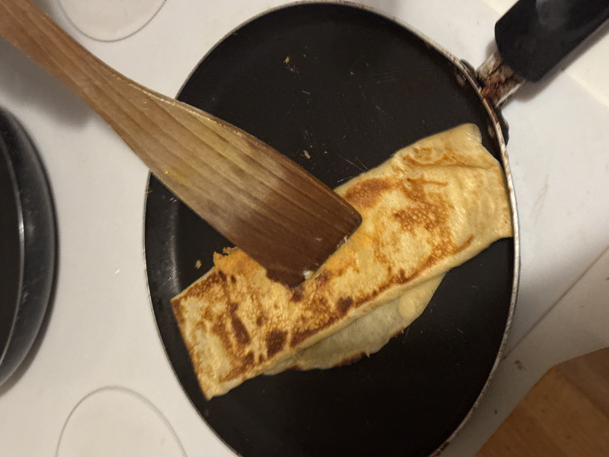 A close-up, overhead shot shows a wooden spatula lifting a folded crepe or pancake in a black non-stick frying pan. The crepe is golden brown with darker browned spots, indicating it is being cooked. The frying pan rests on a white electric stovetop, with a portion of another pan visible in the upper left corner. The background is slightly out of focus, but the stovetop and the kitchen counter below are discernible. The scene appears to be set in a domestic kitchen environment. There are no visible people or specific landmarks that would indicate a location in Budva, Montenegro.