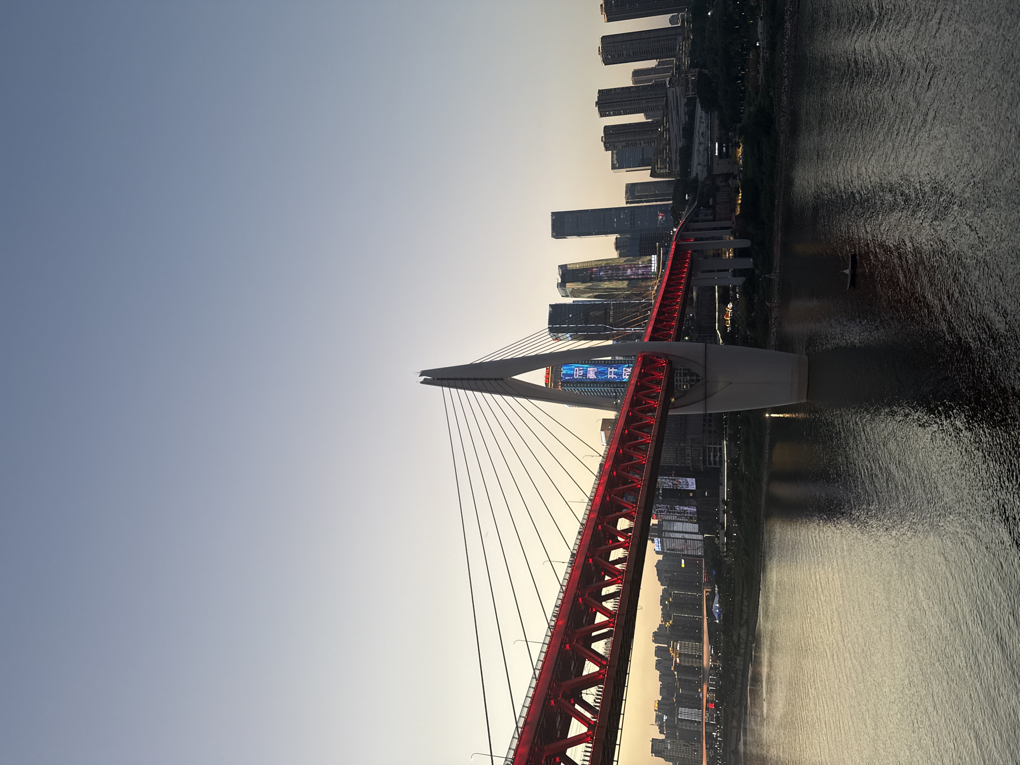 The image shows a modern suspension bridge illuminated with red lights at sunset. The bridge spans a wide river, with a city skyline of tall buildings visible in the background. The sky is a gradient of pale blue and soft orange. The river's surface is dark and reflective, showing ripples. On the bridge, silhouettes of people can be seen walking along the pathway. In the river, a small boat with a single person is visible. The cityscape features a mix of contemporary high-rise buildings, some of which are illuminated with blue and white lights. The overall scene depicts a vibrant urban environment at dusk. The location is Yuzhong District, China.