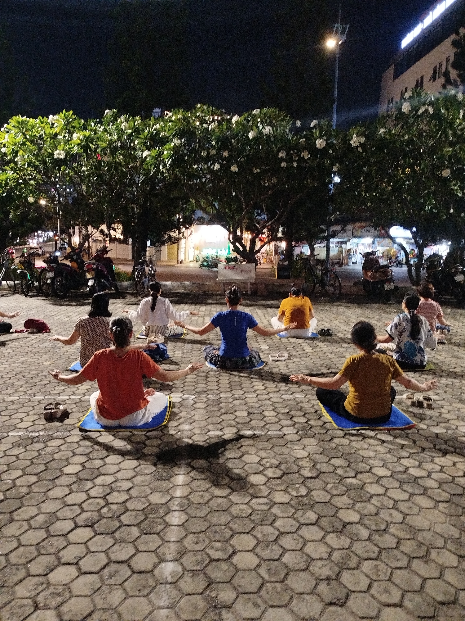 A group of approximately 8 people are sitting in a circle on yoga mats on a paved area, likely a public square or park in Ho Chi Minh City, Vietnam. They are engaged in an activity that appears to be yoga or meditation, with arms outstretched. The setting is at night, illuminated by streetlights and the glow of nearby buildings. The paved surface consists of interlocking hexagonal stones. Trees with white blossoms are in the background, and motorbikes are parked along the edge of the area.