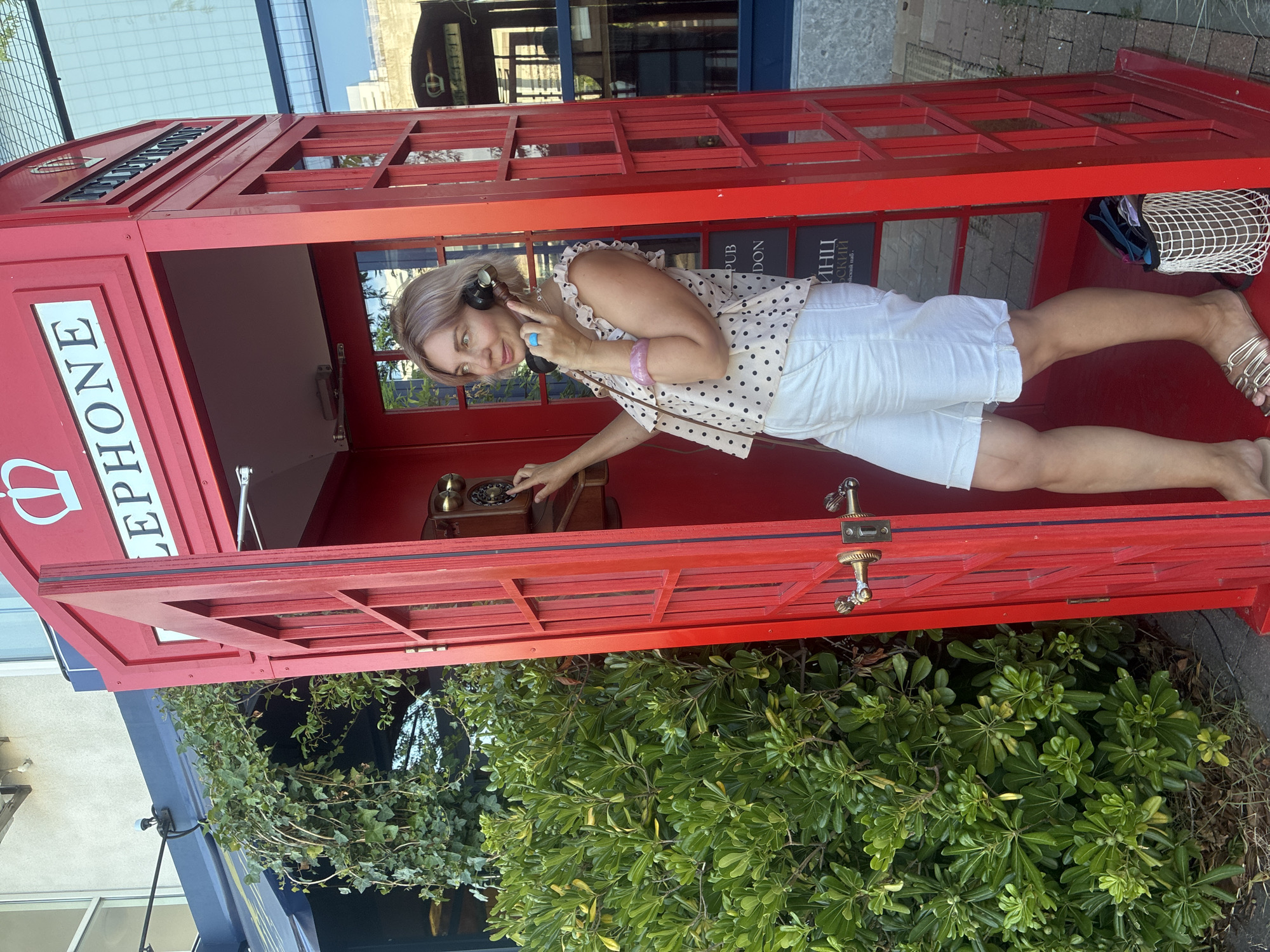 A blonde woman with light makeup is standing inside a red British telephone booth. She is wearing a polka-dot sleeveless top and white shorts, and gold flat sandals. She has a headset on, with the earpiece over her right ear and the microphone near her mouth, and her right hand is on the rotary dial of the telephone. She is looking to her left, with her lips slightly parted as if speaking. She is wearing a large turquoise ring on her right index finger and a pink bracelet on her left wrist. The telephone booth is on a paved walkway, with a lush green bush to the left and a building with large windows to the right. The phone booth has glass panes and a red door that is ajar. A white mesh bag with items inside is on the floor of the booth, near her feet. On the inside of the booth, there are signs that read "PUB LONDON" and what appears to be Russian text. The overall lighting suggests it is daytime. The location is identified as the city of Sirius, Russia.