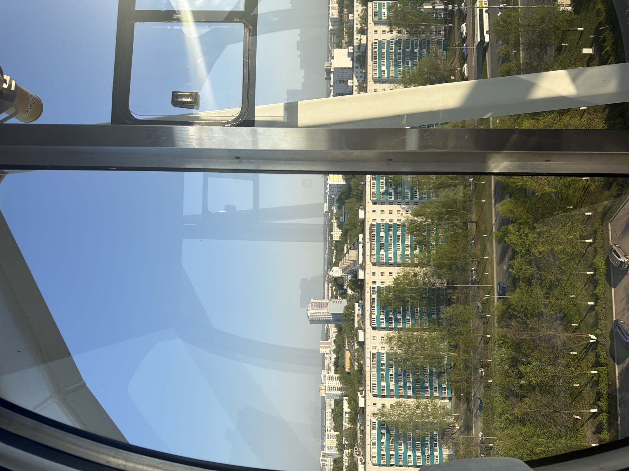 The image shows an urban view from an elevated position, likely a Ferris wheel, in Naberezhnye Chelny, Russia. The foreground consists of the window frame and structure of the viewing cabin, reflecting a clear blue sky. The background displays a dense residential area with numerous high-rise apartment buildings, characterized by a repetitive architectural style with teal accents on the balconies. Rows of trees line the streets between the buildings. Cars are visible parked along the streets. The atmosphere appears bright and sunny.