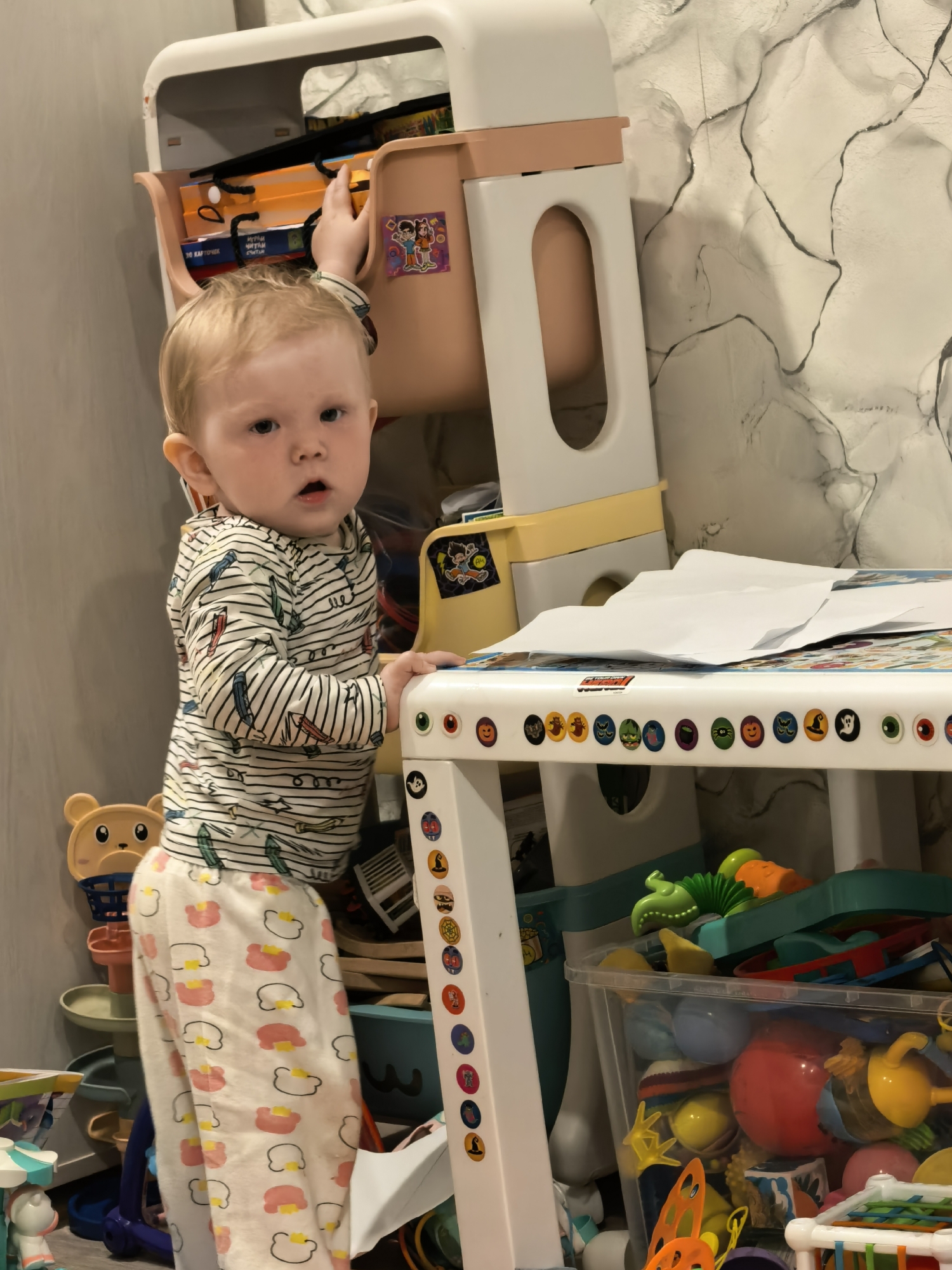 A fair-skinned, blonde-haired baby stands next to a white table with Halloween-themed stickers and sheets of white paper on top, looking towards the viewer. The child is wearing a striped shirt with feather-like accents and white pants with pink cloud-like patterns. The baby is near a pink and white shelf unit with objects within the shelves, and a clear plastic container filled with toys sits beneath the table. The location is identifiable as a domestic indoor space. There is no verifiable evidence to confirm this image was taken in Sterlitamak, Russia.