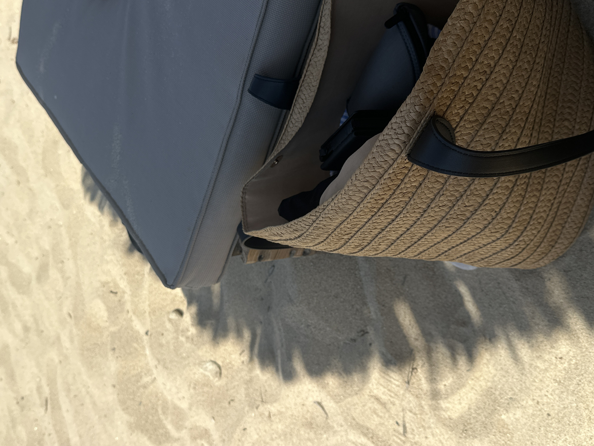 The image shows a close-up view of a straw beach bag with black leather handles, placed on the sand next to a dark gray cushion. The bag contains several dark-colored items, possibly clothing or accessories. The sand is light brown and shows some texture and small pebbles. Shadows of leaves are cast across the sand, indicating the presence of nearby vegetation, possibly from trees. In the background, part of a cushioned lounge chair is visible. The scene suggests a relaxed setting at a beach, likely Ofrynio Beach in Greece, given the context. There are no people visible in the image.
