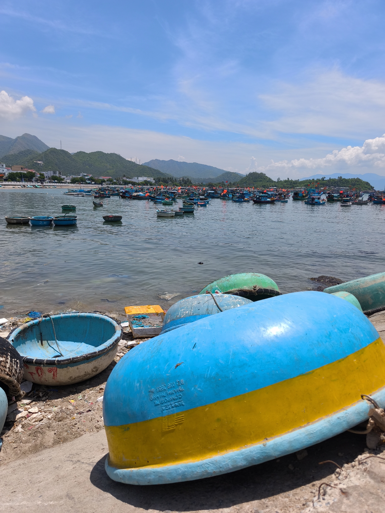 The image depicts a harbor scene in Vĩnh Lương, Vietnam, under a clear blue sky with scattered clouds. In the foreground, several boat hulls are visible on the shore. The largest, a bright blue and yellow boat, is overturned and takes up a significant portion of the frame, with text partially visible on its side. To the left, a smaller, blue, round boat is also present, with a white rim and red markings. Debris litters the shore around these boats, including plastic fragments and other refuse.
The middle ground shows a body of water filled with numerous boats of various sizes and colors, mostly in shades of blue and some with red flags. These boats appear to be docked or anchored in a calm bay. Some are smaller, round fishing boats, while others are larger, more modern vessels.
In the background, a coastal town stretches along the shoreline, with buildings nestled at the base of green, mountainous hills. The hills are lush with vegetation. The overall impression is a busy fishing harbor in a coastal community. There are no visible people in the frame.