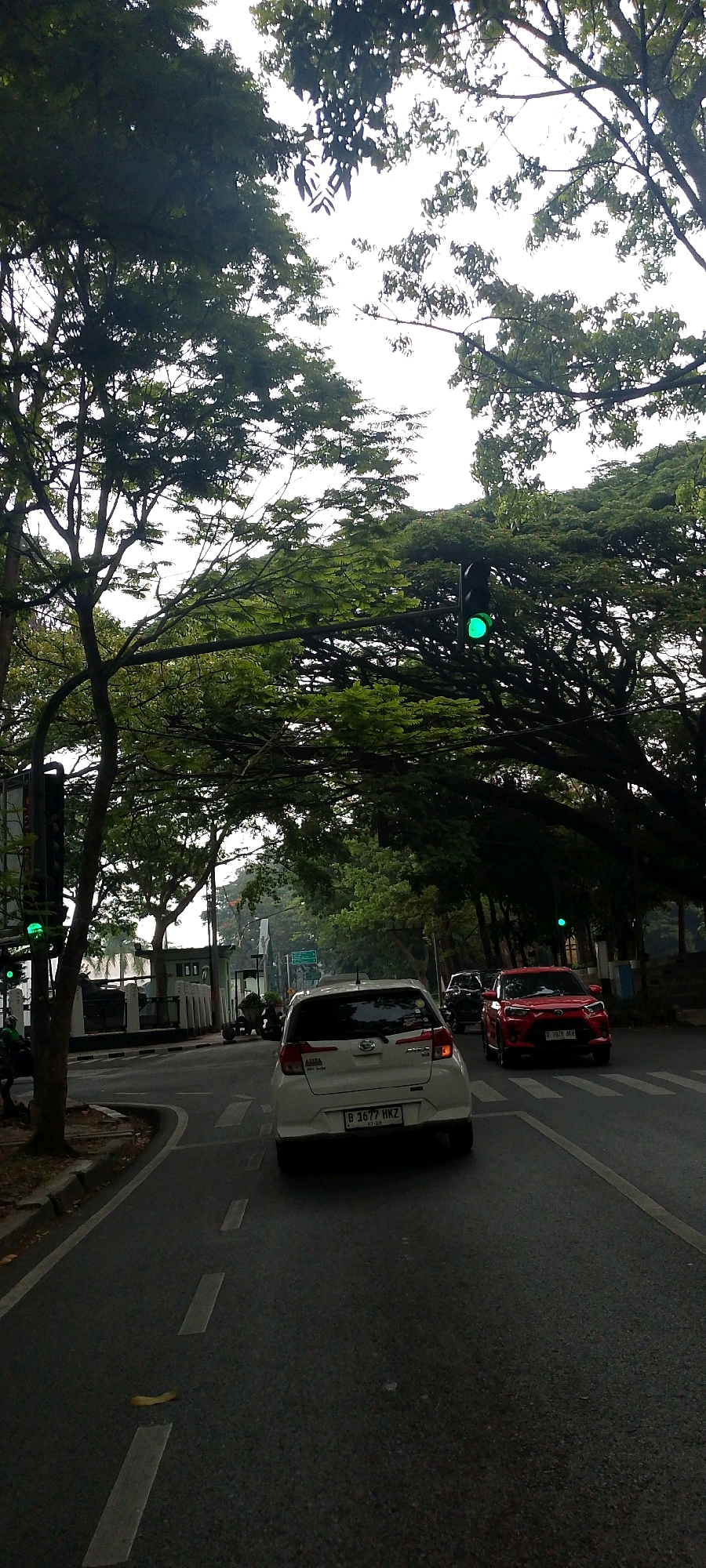 The image shows a street scene in Bandung City, Indonesia. A white car with license plate B 1677 HKZ is in the foreground, driving on a road lined with trees. A red car is in the lane next to it, also driving. Traffic lights are visible, with the green light illuminated. The sky is overcast, and the trees are full of green leaves, suggesting it is daytime.