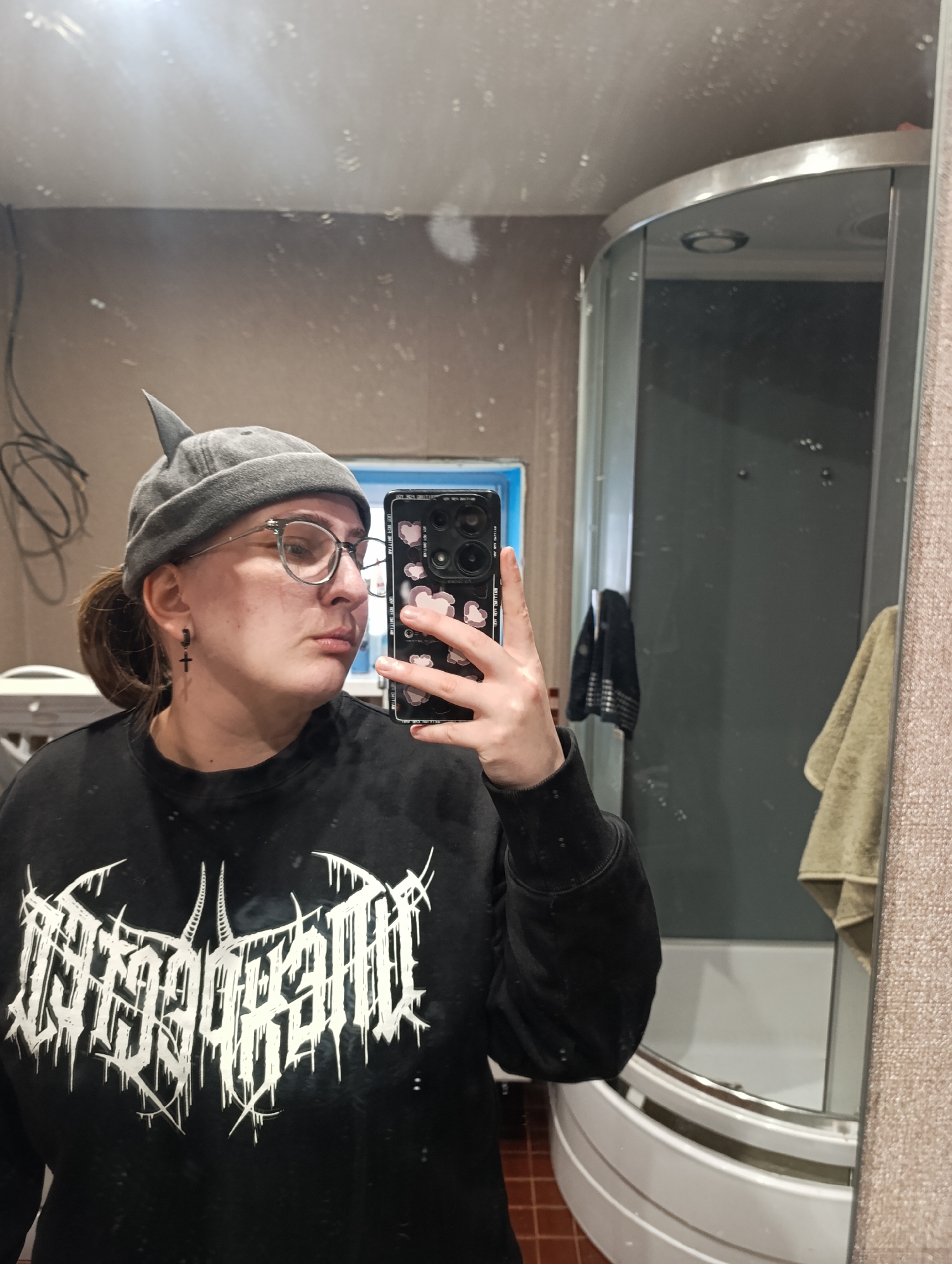 A person with round glasses and a gray peaked beanie is taking a selfie in a bathroom. The person is wearing a black sweater with white gothic style lettering. The phone case has a pattern of pink hearts. A black cross earring is visible on the left ear. The bathroom features a mirror, a shower stall, and a brown wall. A partially visible washing machine is in the background to the left. Based on the presence of Russian text, the location is likely Chaikovsky, Russia.