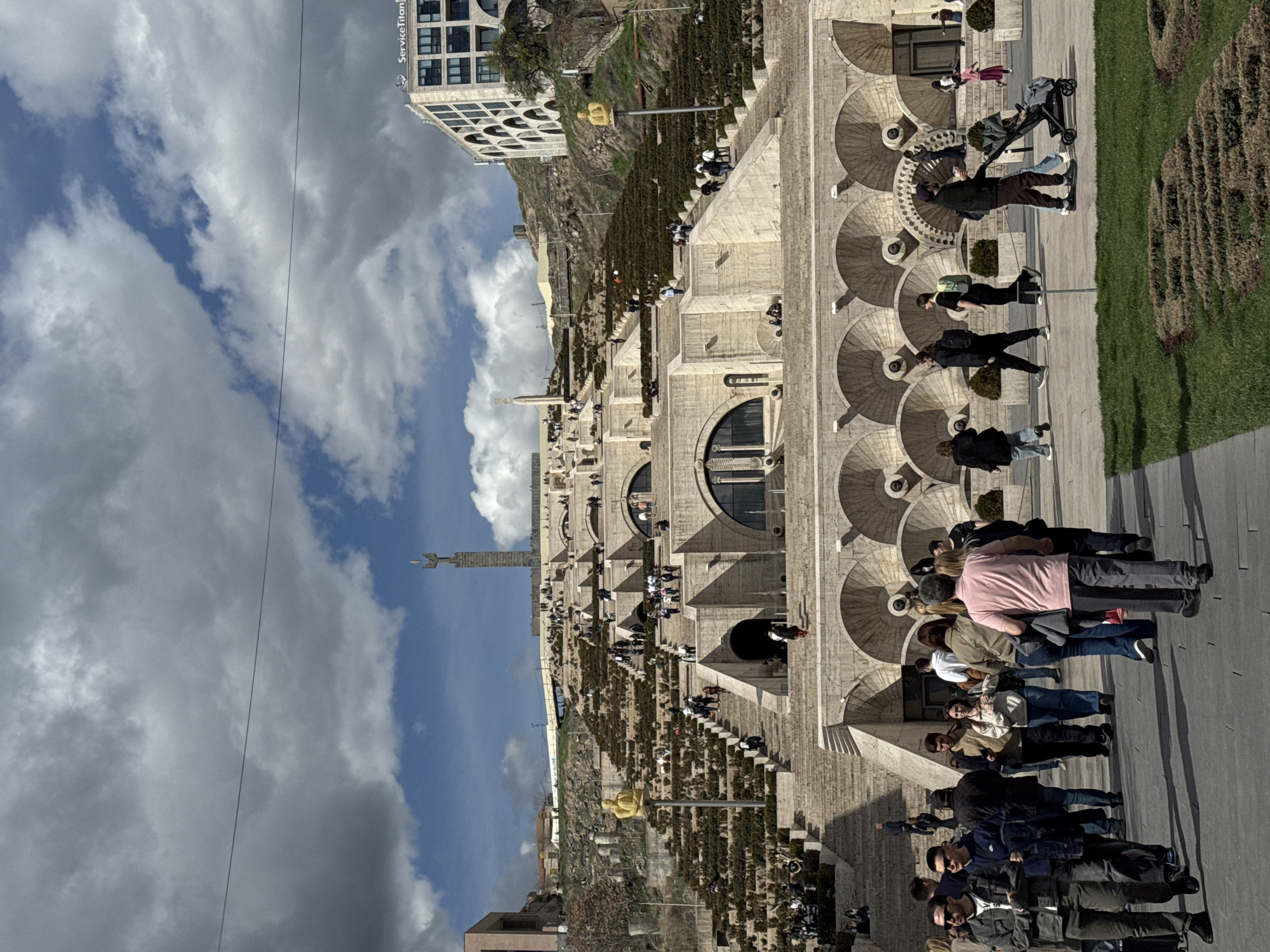 The image depicts the Cascade, a monumental stairway in Yerevan, Armenia. The structure is made of light-colored stone and features multiple levels adorned with fountains and sculptures. People are walking along the staircase and the adjacent paved areas. The sky is visible with varying cloud cover. A modern building stands adjacent to the Cascade. A golden statue is present on the steps leading up the Cascade.