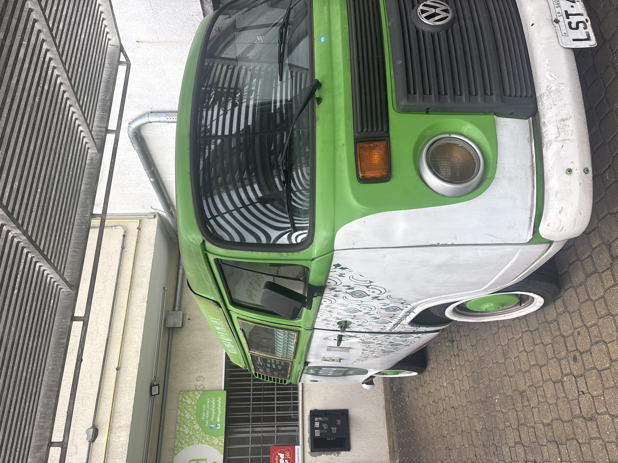 A green and white Volkswagen Kombi is parked on a brick-paved surface in front of a building with a white facade and a metal awning. The Kombi has white wheels with green hubcaps. The side of the van features a repeating green organic pattern. A sign on the wall to the left of the van reads "Siga-nos nas redes sociais" followed by social media handles and a QR code. The front of the Kombi has a large round headlight, a silver Volkswagen emblem, and a license plate that reads "LST-4... SP - SAO PAULO". The building appears to be a commercial establishment, possibly a food truck or a small business. The overall setting suggests an urban environment in São Paulo, Brazil.