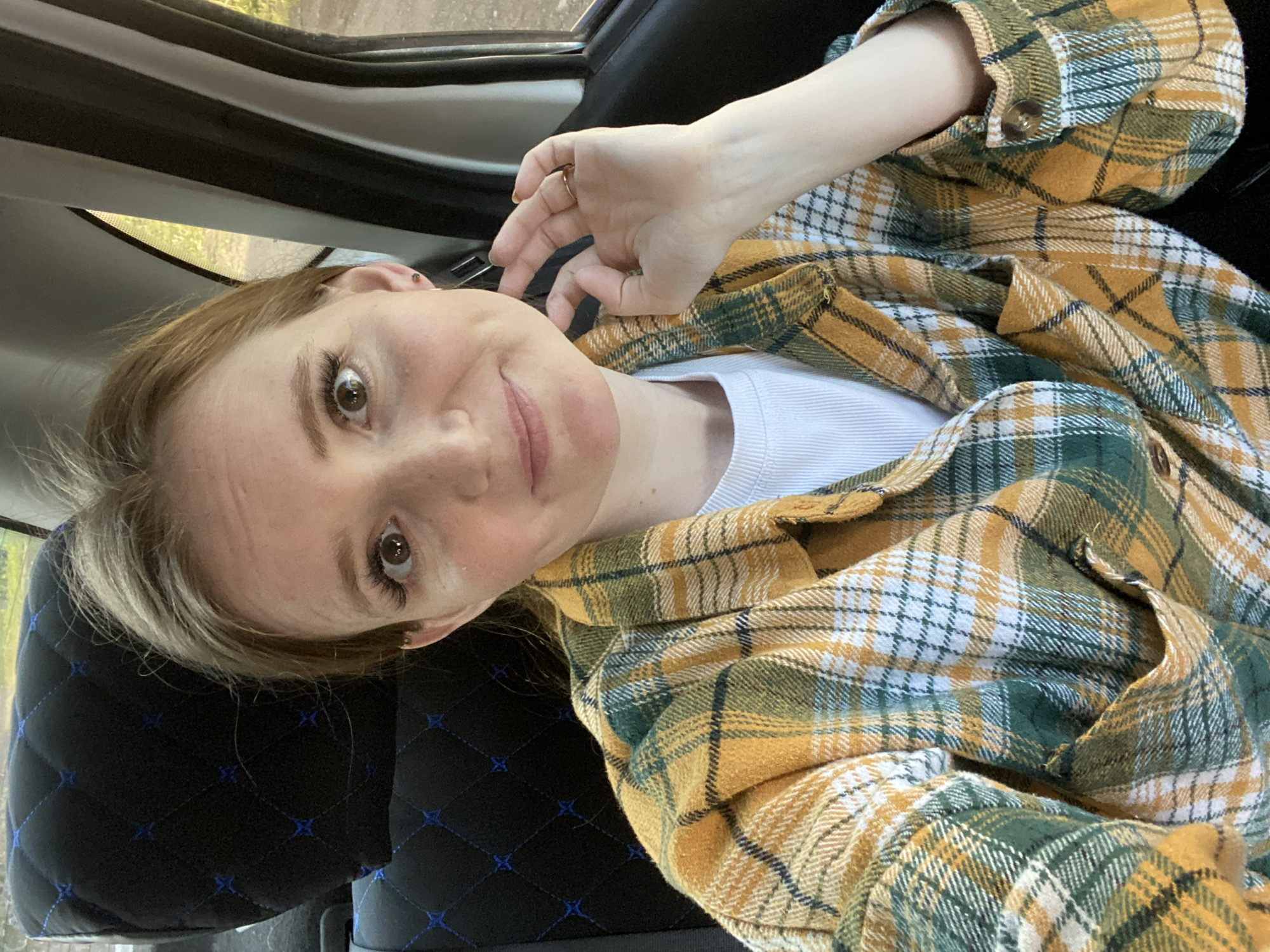 The image captures a selfie of a young-looking individual inside a vehicle, in Chaikovsky, Russia. The person, likely female, has light brown hair with blonde highlights, brown eyes, and fair skin. They are looking directly at the camera with a slight, friendly smile.
The individual is wearing a white ribbed t-shirt, over which an open plaid button-up shirt is donned. The plaid pattern features prominent colors of mustard yellow, dark green, and white, with thin dark lines outlining the squares. A golden ring is visible on the ring finger of their right hand, which is raised near their face, appearing to hold a phone to take the picture or adjust their hair.
The setting is the interior of a car, with a black seat featuring a distinctive quilted or stitched pattern in blue visible behind and beside the person. The upper part of the car's door frame and a window are partially visible in the background, showing blurred greenery and suggesting it is daytime. The overall impression is casual and relaxed. There is no visible text in the image.