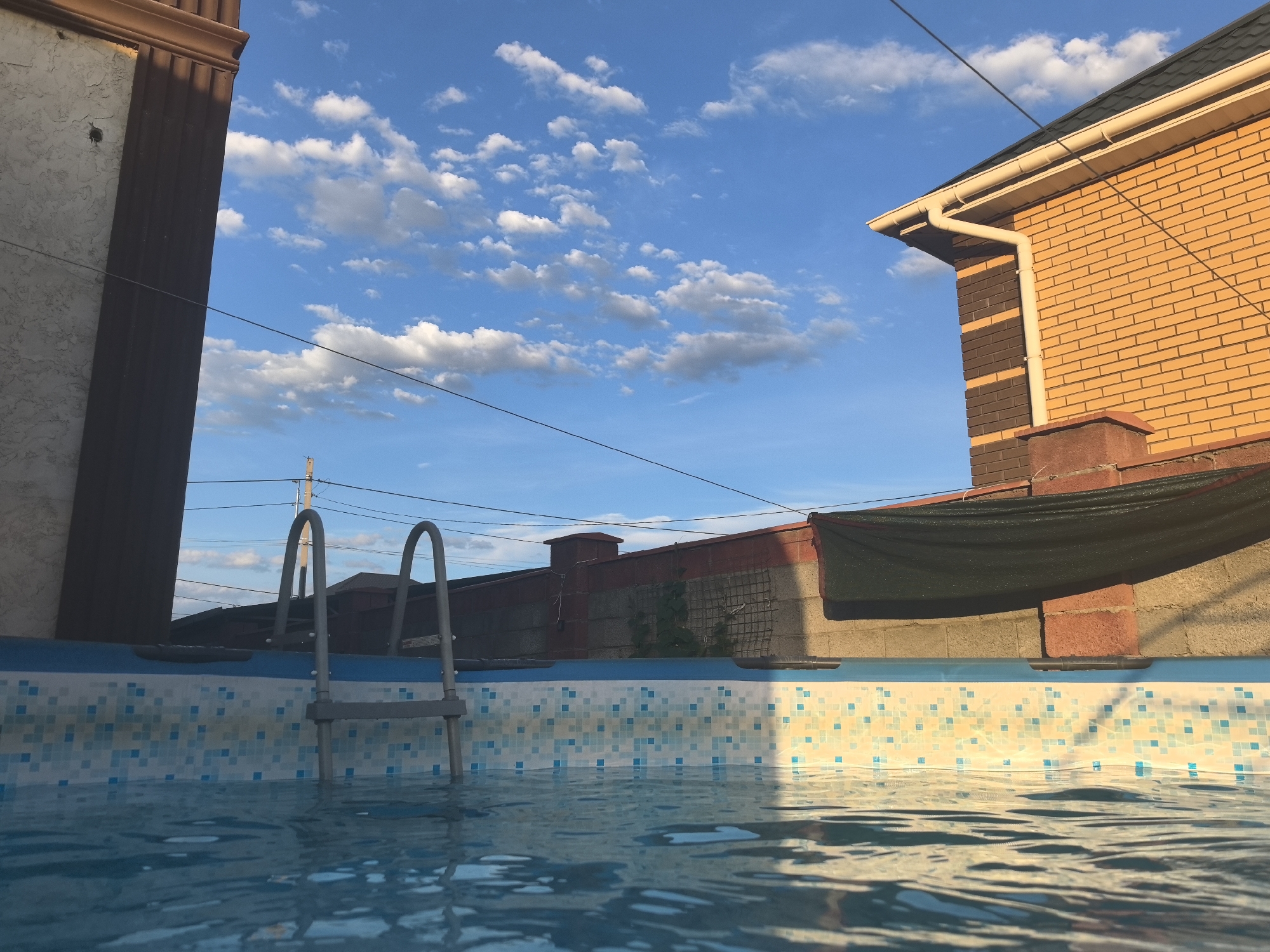 The image captures an outdoor scene, likely a backyard in Taldykorgan, Kazakhstan. In the foreground, the rippling surface of a swimming pool fills the bottom half of the frame, its light blue water reflecting the sky. The pool walls are lined with small, colorful tiles, predominantly white with scattered blue and teal squares. A silver metal ladder with arched handrails emerges from the pool on the left side, its base submerged in the water.
The background reveals the edges of structures. On the far left, a portion of a building with textured beige siding and a dark brown, fluted architectural element is visible. To the right, a bright orange brick building with a dark grey shingled roof and a white gutter system dominates the upper right corner. A red brick chimney or decorative pillar is visible in front of this building.
A dark green mesh screen is stretched across the top of a brick wall behind the pool, casting a shadow. The sky above is a vibrant blue, dotted with fluffy white clouds that are beginning to catch the warm light of the setting sun. Power lines crisscross the sky, and a utility pole is visible in the distance.
The time of day appears to be late afternoon or early evening, indicated by the soft, warm light and the long shadows cast by the structures. The weather seems pleasant, with a clear blue sky and scattered clouds suggesting fair conditions. The overall mood is tranquil and inviting, evoking a sense of leisure and relaxation associated with a poolside setting. No text is visible in the image.