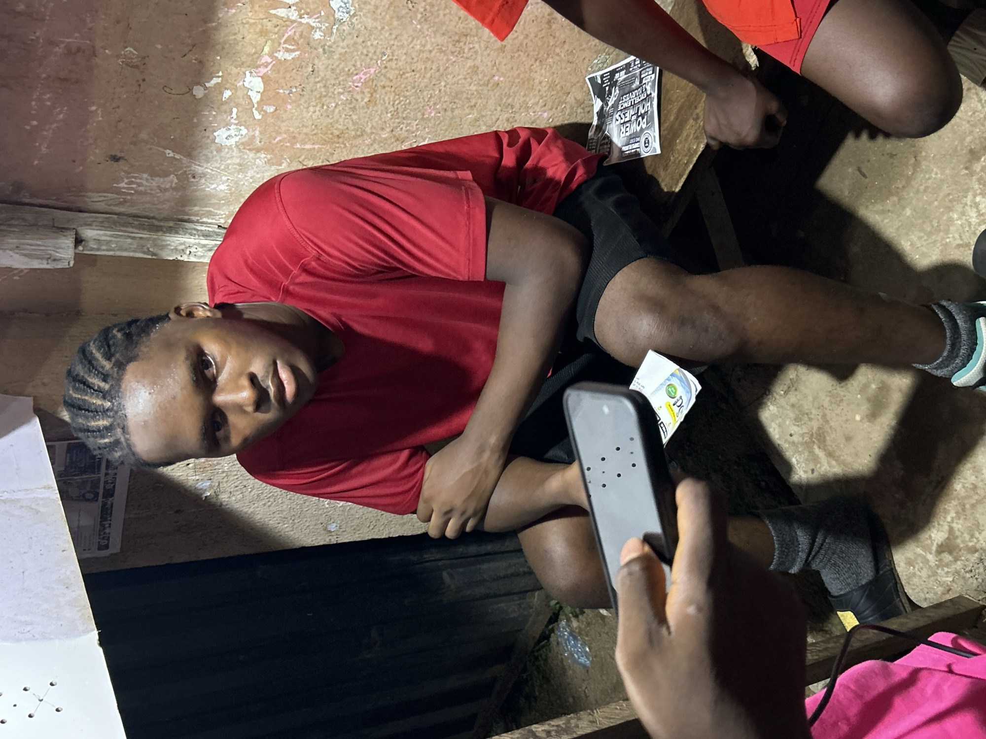 A young man with braided hair, wearing a red shirt and black shorts, is seated on a rough surface within a dilapidated structure in Ihiagwa, Nigeria. He appears to be looking directly at a mobile phone being held in front of him. A second person, only partially visible, is wearing a red shirt and shorts. The environment is poorly lit and appears to be a makeshift space. There is printed material on the surface near the seated man and an object with green and white packaging. The floor is bare.