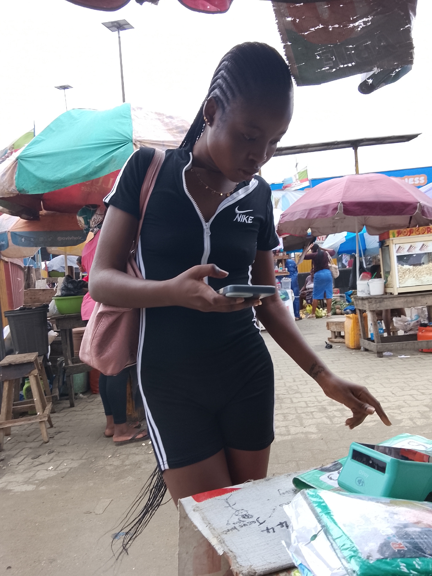 This media file captures a bustling outdoor market scene, likely in Lekki, Nigeria. The central subject is a young Black woman dressed in a black Nike athletic outfit. She is holding a smartphone in her hands, her gaze directed downwards towards it. She also has a pink bag slung over her shoulder. Her hair is styled in braids that cascade down her back.
The background reveals a vibrant market atmosphere with numerous stalls and umbrellas providing shade. Various objects are visible, including plastic containers filled with produce, a popcorn machine, and what appears to be other merchandise on display. Other individuals can be seen in the background, engaged in their own activities, contributing to the lively environment. One woman in a pink garment and headscarf stands near a stall, while another woman in a blue skirt is seen further in the distance.
The setting suggests it is daytime, as indicated by the ambient light. The sky appears bright, suggesting fair weather. The ground is paved with bricks. The presence of multiple stalls and the general setup strongly indicate a marketplace. The woman's focused expression on her phone might suggest she is communicating, browsing, or perhaps making a purchase, though her hand gesture also hints at interaction with an item on a stall. No specific emotions are overtly displayed, but the overall scene conveys a sense of everyday commerce and activity.
Visible text includes the "NIKE" logo on the woman's outfit. Additionally, the word "POPCORN" is clearly visible on the popcorn machine. A sign with the word "ESS" is also partially visible in the background, possibly part of a brand name.