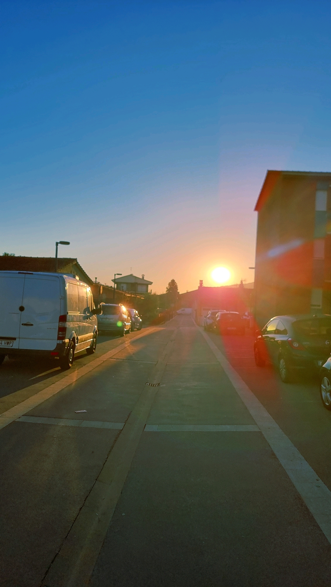 This is a vertical, eye-level, outdoor, daytime photograph taken at street level in Sant Joan Les Fonts, Spain. The sun is setting, casting a warm orange glow and lens flares across the scene. The sky is a clear, bright blue. In the foreground, a paved street runs towards the horizon, with white lines marking lanes or parking spaces. On the left side of the street, a white cargo van is parked. Several other cars are parked along the right side of the street, including a dark-colored hatchback in the immediate foreground. In the middle ground, there are more cars parked on both sides of the street, and a few residential buildings are visible. One building on the right is a dark, multi-story structure with a strong lens flare coming from its side. Further in the distance, more houses and a distant tree can be seen. The overall impression is a quiet, suburban street scene at dusk.