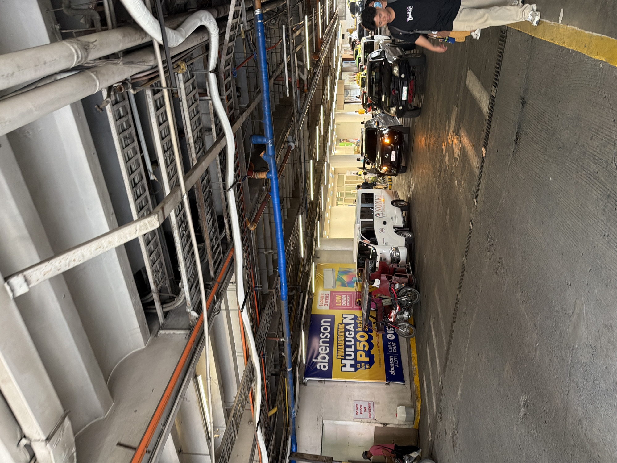 The image depicts a street scene in what appears to be a covered parking area or a ferry terminal, likely in Davao City, Philippines, given the presence of a tricycle and a tri-star license plate visible on one of the vehicles. Overhead, a complex network of pipes, conduits, and structural beams is visible, suggesting an industrial or transit infrastructure.
In the foreground and midground, several vehicles are present, including a white van, a red and black tricycle, a black SUV, and other cars. The vehicles are in a line, suggesting traffic or a waiting area. One white van has "NONKI" written on its side.
A large yellow and blue banner is prominently displayed on the left side of the image, featuring the "abenson" logo and text in Filipino, promoting a product or service with "Hulugan P50 kada". Below the banner, a small white sign reads "DO NOT BLOCK THE DRIVEWAY".
Several people are visible in the scene. On the far left, a person in a pink shirt is standing near a wall. In the midground, a person is walking towards the right, wearing a black t-shirt and khaki pants. Other individuals are seen near the vehicles, some appearing to be drivers or passengers.
The ground is paved with concrete, and yellow lines mark lanes or boundaries. There are also some drains or grates visible in the pavement. The lighting suggests an overcast day or an indoor environment with artificial lighting. The overall impression is that of a functional, busy area, possibly related to transportation or retail.