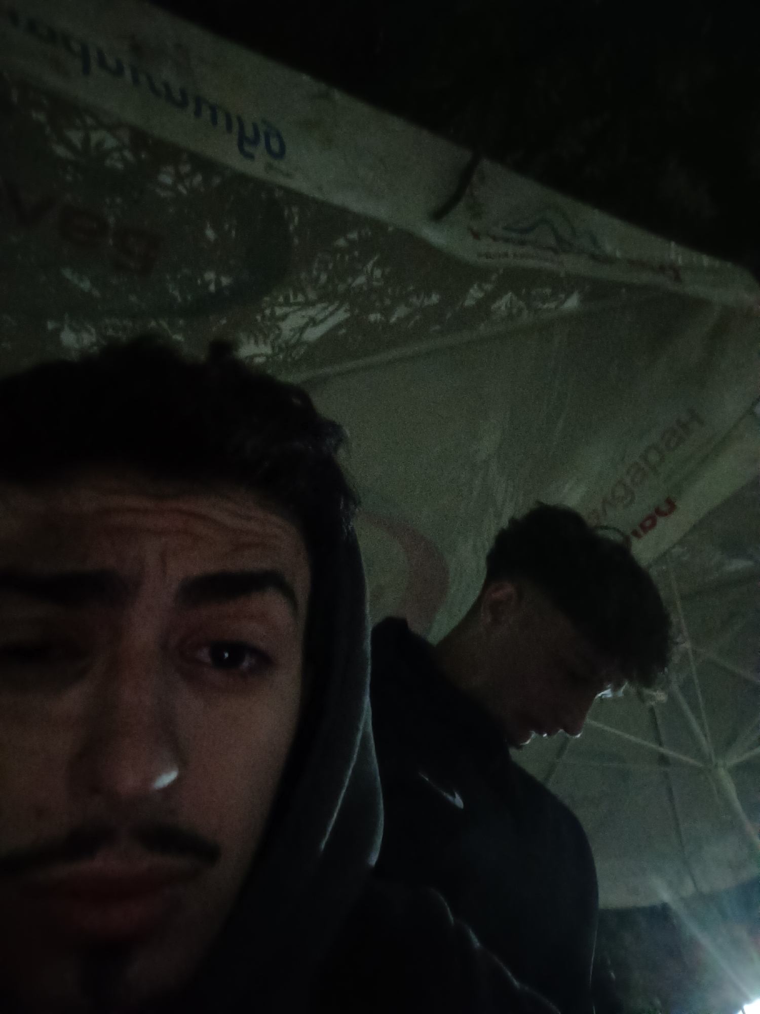 Two young men are under a white umbrella with writing on it, including "HadebgapaH." The image is dark and poorly lit, with the man in the foreground facing the camera, while the other man is in the background, looking down. The location is likely in Pripek, Bulgaria.