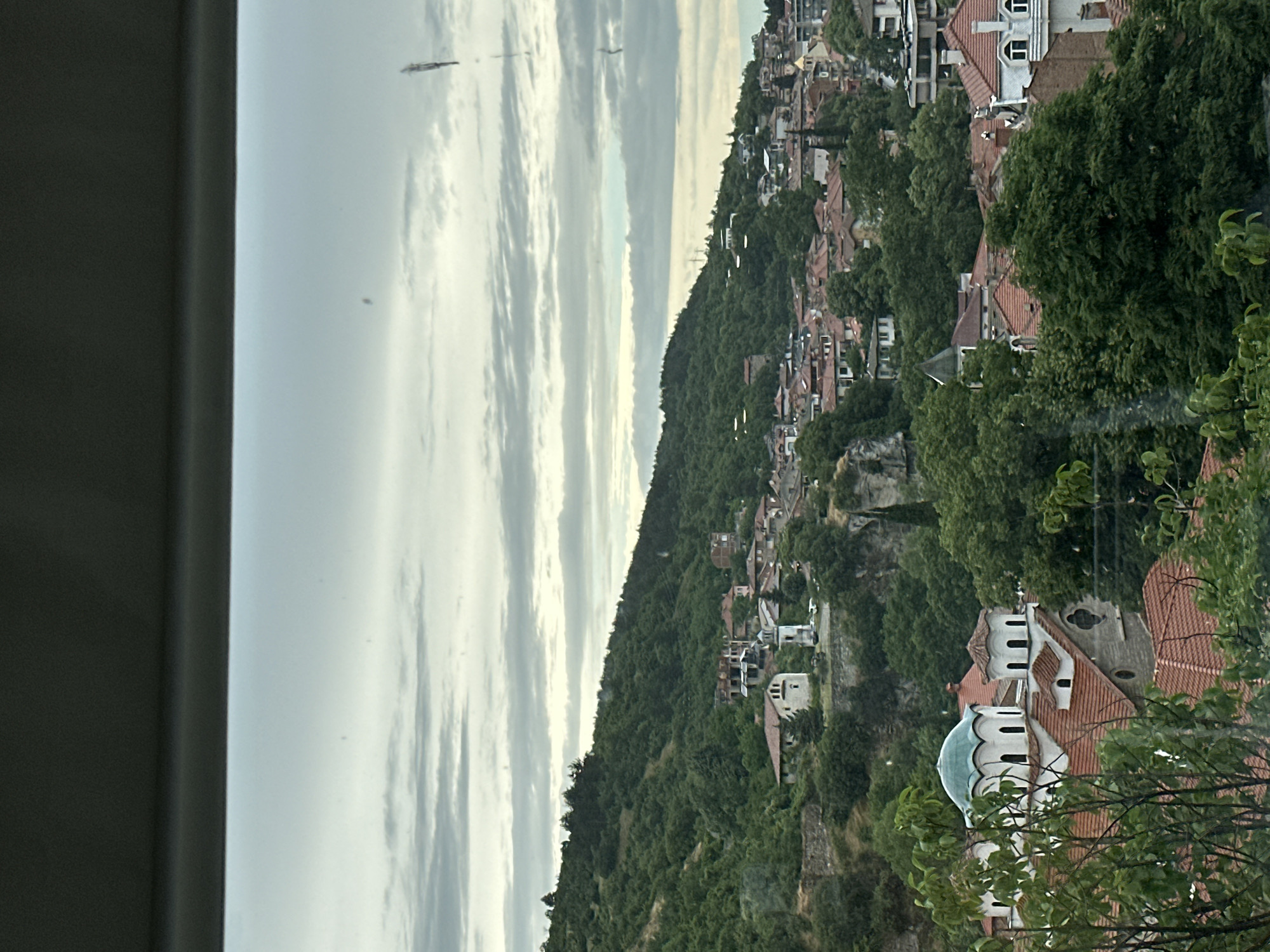 This is a wide, exterior, eye-level, landscape shot of Asenovgrad, Bulgaria, taken on an overcast day. The image is framed from a vehicle, with the top of a car's dashboard visible in the foreground. The scene depicts a densely populated hillside town, characterized by its red-tiled roofs and lush green trees.
In the foreground, a prominent church with a light blue dome and white walls stands out amidst the foliage. This church appears to be of Byzantine or Orthodox architectural style, featuring arched windows and smaller domes. Further into the scene, the town sprawls across the hillside, with a variety of buildings displaying traditional red-tiled roofs. Some structures show signs of age and disrepair, while others appear more modern. A steep, tree-covered hill rises behind the town, dominating the upper portion of the image. The sky is filled with heavy, grey clouds, creating a muted and diffused light. There are no people or animals visible in the frame. The overall impression is one of a historic European town nestled into a verdant, hilly landscape.