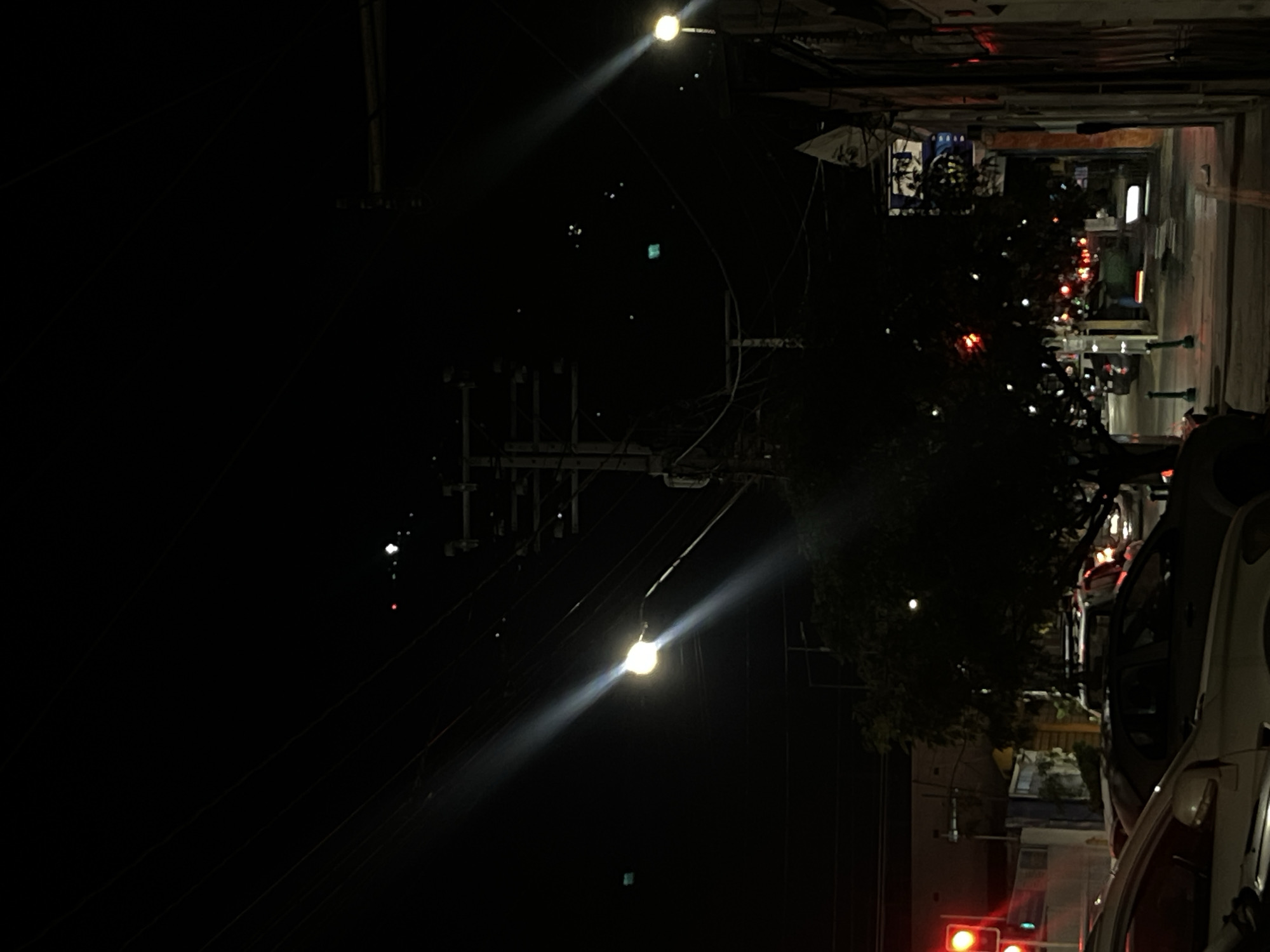 The image depicts an urban street scene at night. The main illumination comes from streetlights that cast bright beams of light. Power lines crisscross the sky. On the right side of the image, there's a parked car and blurred figures of people walking on the sidewalk. Further down the street, there are faint lights suggesting shops or buildings. A traffic light with red lights is visible. The scene is located in Unknown, Unknown.