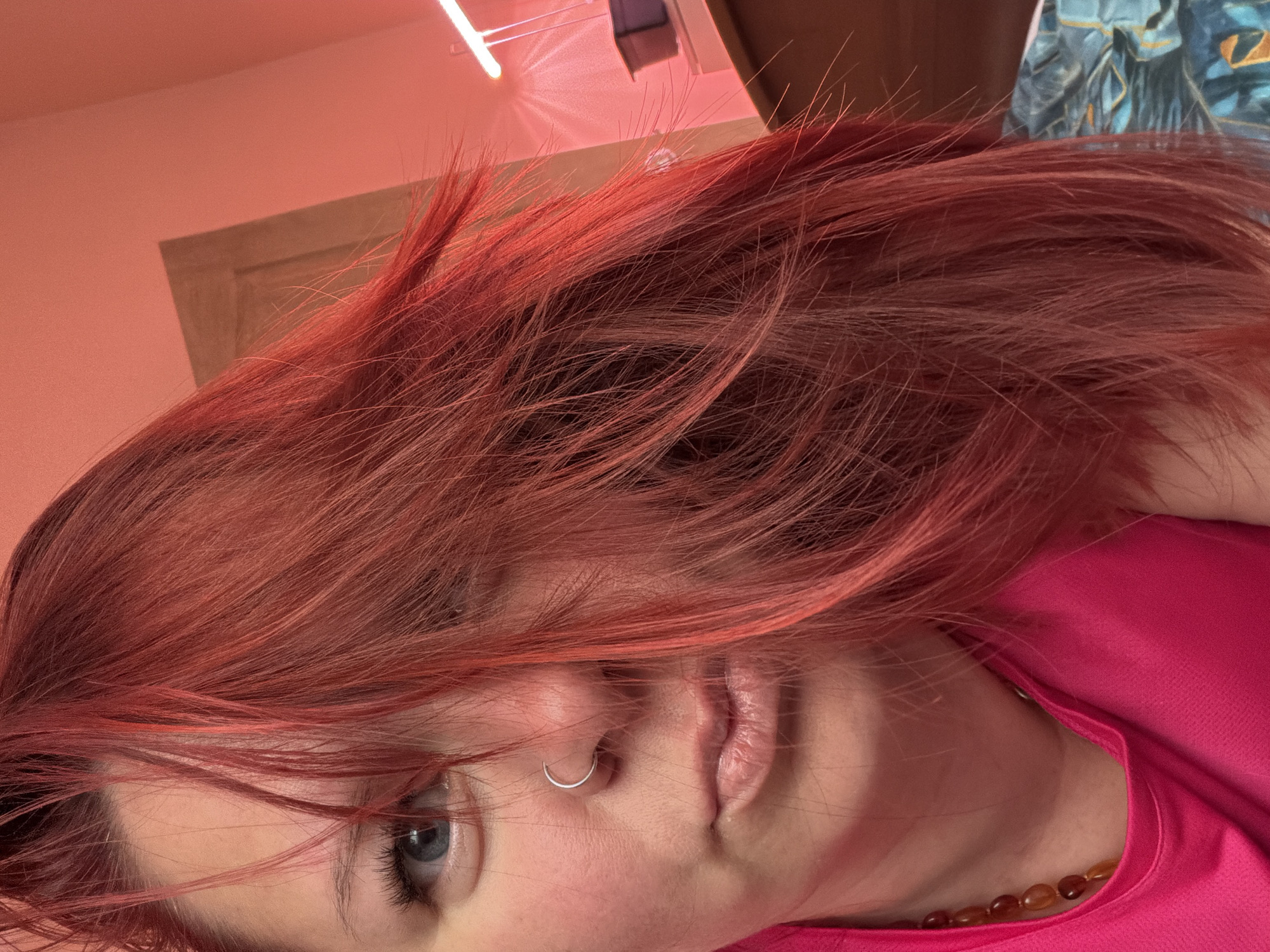A close-up, angled portrait depicts a person with red hair, fair skin, and blue eyes. They are wearing a pink shirt and a beaded necklace with amber-colored beads. A silver nose ring is visible. The background includes a beige wall, a light fixture emitting a red glow, and a partially visible piece of artwork or fabric with a blue pattern. There is no specific evidence or indication to suggest this photograph was taken in Chaikovsky, Russia. The location is unknown.