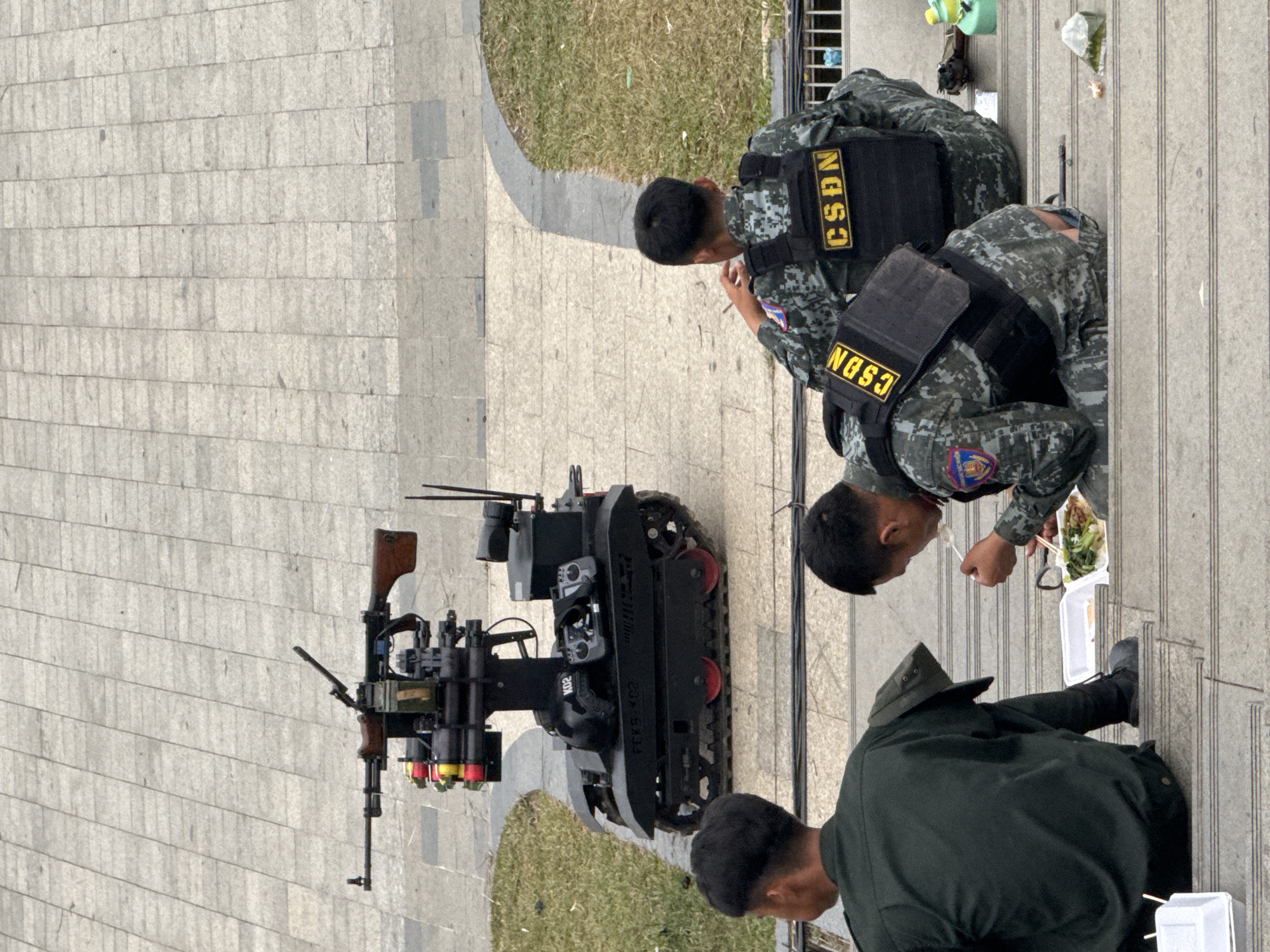 In Da Lat, Vietnam, three individuals in military-style uniforms are seated on concrete steps. A robotic device, armed with a machine gun and what appear to be rocket-propelled grenades, is positioned on a paved surface behind them. The individuals are engaged in eating. One is actively consuming food from a white container with chopsticks. The robotic device has treads and is painted black. The environment suggests an outdoor, possibly public, area with a grassy patch and paving stones visible. The overall scene conveys a sense of security or observation.