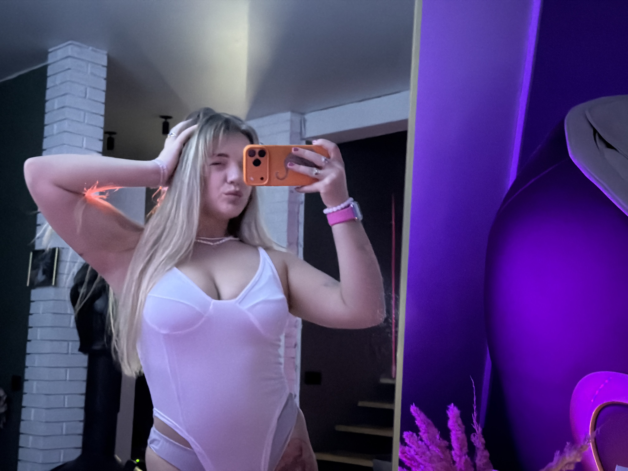 The image is a mirror selfie of a young woman indoors. She is wearing a light-colored bodysuit and holding an orange phone with a triple-lens camera in her right hand. She is winking with her left eye and has a slight pout. She has blonde hair and is wearing a pearl necklace and a pink watch or bracelet on her right wrist. Her left hand is also raised towards her head.
Behind her, there is a brick-like design element on the wall, and a dark object resembling a coat or statue is partially visible. In the mirror reflection, a staircase is faintly seen. To the right, another structure with purple lighting and decorative plants are seen, suggesting the presence of more items.
The location of the image is potentially in the city of Большая Шильна, Russia.