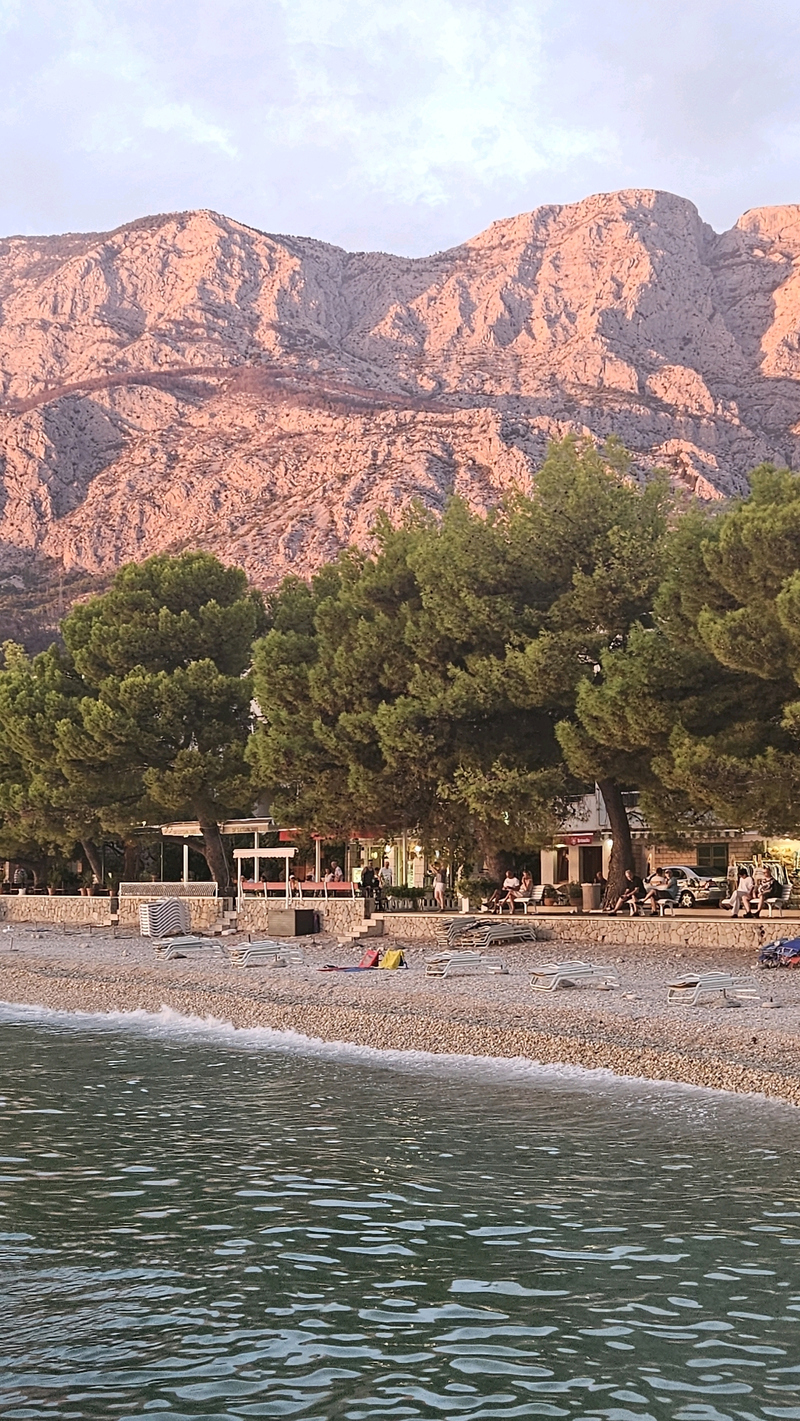 The image depicts a beach scene in Općina Tučepi, Croatia, with a rocky mountain range in the background illuminated by the warm glow of sunset. The foreground is dominated by the calm, rippling water of the sea. The beach itself is covered in small, light-colored pebbles, with a line of neatly stacked white lounge chairs and umbrellas. Further back, a paved walkway runs parallel to the beach, lined with more lounge chairs and a few scattered individuals. Several trees with lush green foliage form a natural barrier between the beach and structures that appear to be cafes or small shops. Some people are seated at tables outside these establishments, and a parked car is visible. The overall atmosphere is serene, suggesting a peaceful evening at a coastal resort.