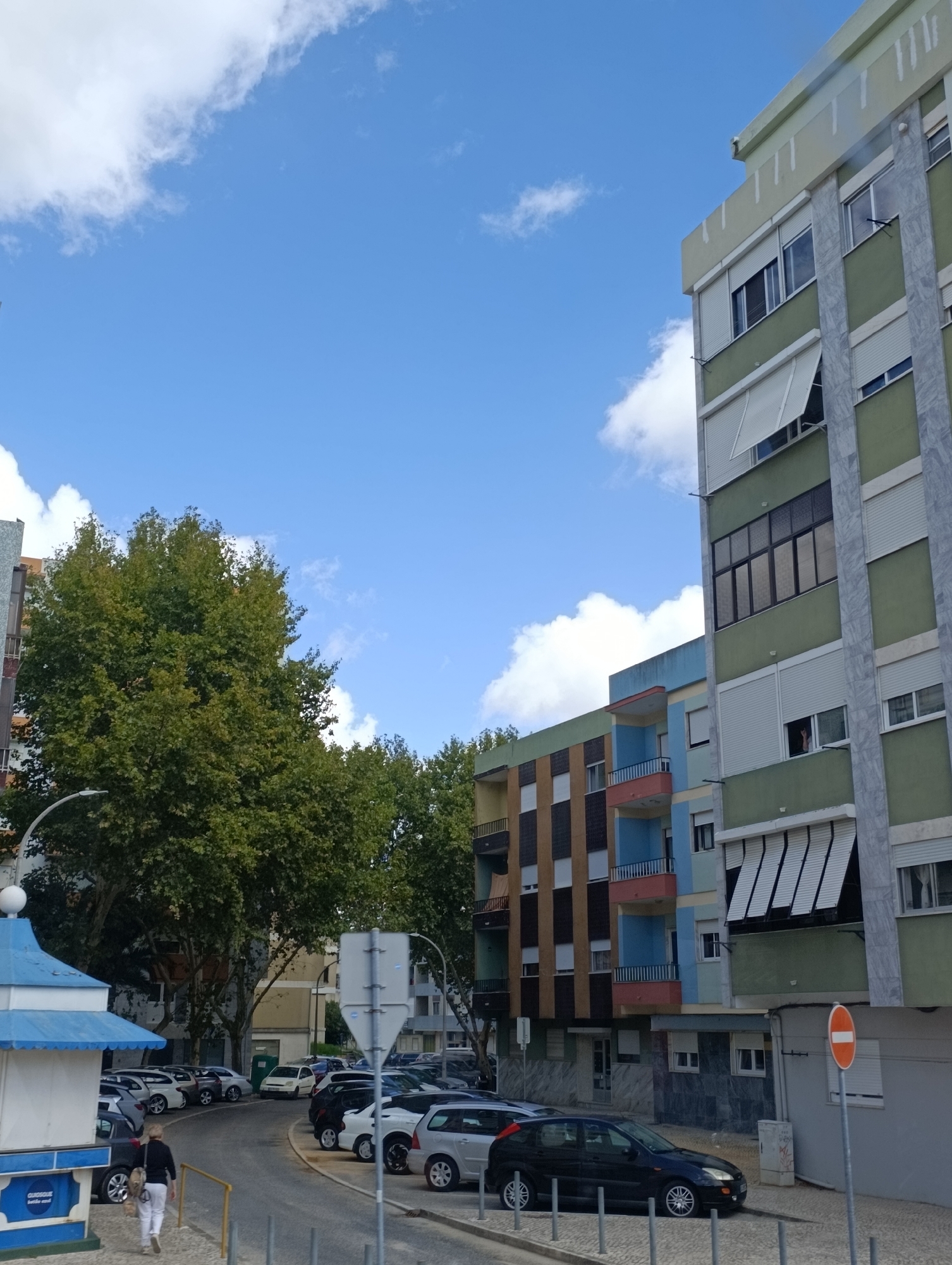 In Setúbal, Portugal, a street scene unfolds under a partly cloudy sky. On the right, a multi-story building, primarily painted in a muted green with marble-patterned accents, stands tall. It features multiple windows, some with white roll-down shutters, and balconies. To the left of this building, another apartment complex displays a more vibrant facade, with sections painted in shades of blue, yellow, and brown, each with small balconies. A large, leafy green tree dominates the mid-left of the frame, partially obscuring the buildings behind it.
In the foreground, a paved road curves from the left, bordered by parked cars. Several vehicles, including a black hatchback, a white sedan, and a silver hatchback, are visible. A woman in a dark top and white pants, carrying a beige backpack, walks away from the camera on the left side of the road. A blue and white kiosk-like structure is positioned in the lower left corner, with some text visible on its side. A street lamp with a curved arm extends over the road from the left. A white sign with a red circle and a horizontal bar, indicating "no entry" or "do not enter," is affixed to a pole on the right side of the street. The environment appears to be an urban residential area.