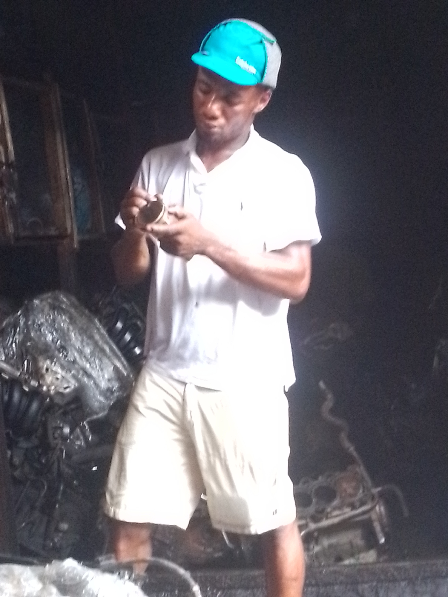 In a dimly lit location, presumably a mechanic's shop in Onitsha, Nigeria, a man wearing a bright blue cap, a white polo shirt, and light-colored shorts is meticulously examining a cylindrical object with both hands. The background is cluttered with car parts, including engines partially covered in plastic wrapping. The overall impression is of an individual engaged in close inspection or repair work within a workspace.