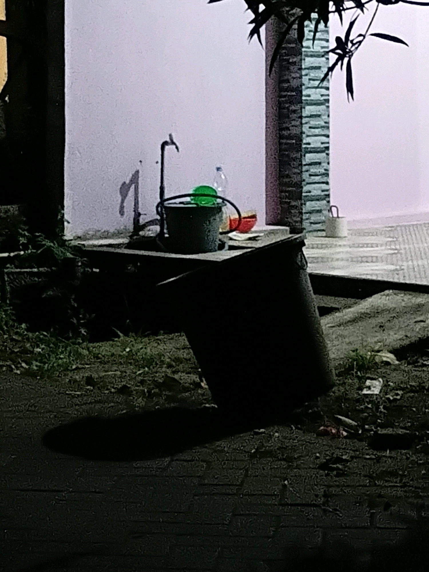 The image depicts an outdoor scene at night in Cikal, Indonesia. In the foreground, a large, dark, cylindrical object is positioned on uneven ground covered with leaves and debris. A distinct shadow of this object is cast onto a paved surface. To the left of this object, more debris and foliage are visible.
In the mid-ground, against a light-colored wall, a water faucet is mounted. A dark bucket with a decorative pattern sits beneath the faucet, containing a bright green object, possibly a bowl or lid. A clear plastic bottle and a red bowl are also present near the bucket. Further to the right, a section of a patterned wall or column is visible, with dark leaves from a tree hanging in the upper right corner of the frame. In the background, a light pink wall forms the backdrop, and a tiled pathway is discernible. A small, light-colored bucket with a handle is situated on this pathway. The overall lighting suggests a low-light or nighttime environment.