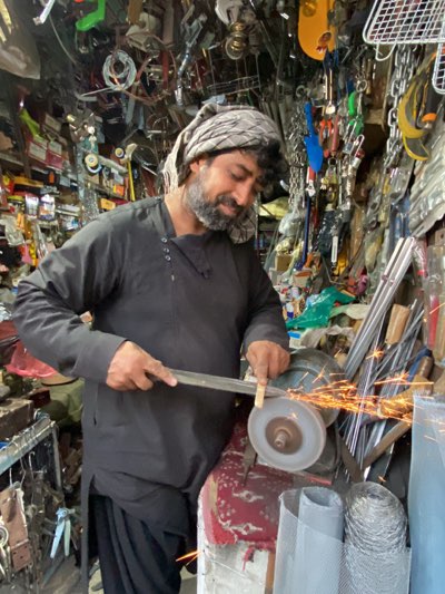 In an overflowing hardware shop in Kandahar, Afghanistan, a middle-aged man with a grey-streaked beard and a patterned turban concentrates on sharpening a metal blade against a spinning grinding wheel. Sparks erupt from the point of contact. The shop is crammed with tools, chains, locks, metal rods, and rolls of wire mesh, hanging from the ceiling and filling every available space. The man is wearing a dark, long-sleeved shirt, and the base of the grinding wheel is painted with red and white patterns.