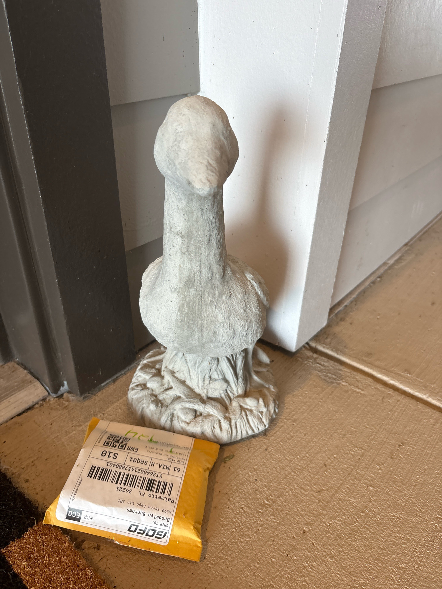 A light gray, stone statue of a stylized goose is positioned on a tan tiled floor next to a dark gray door frame and a white post. To the left of the statue, a yellow, padded envelope is lying on the floor. The envelope has a shipping label with text that includes "Palmetto FL 34221", "Brooklyn Burrows", and "GOFO". The statue is about two feet tall and depicts a goose with a rounded head, an elongated neck, and a bulbous body resting on a textured base. The envelope appears to be a package delivery.