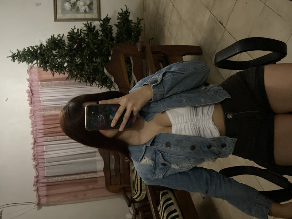 A person sits in a chair, their face obscured by a smartphone. They wear a white top, denim jacket, and black shorts. A small Christmas tree is visible in the background, along with curtains and a framed picture on the wall. The image appears to be taken in a residential setting, with wooden furniture visible behind the chair. The photo is rotated to the side.