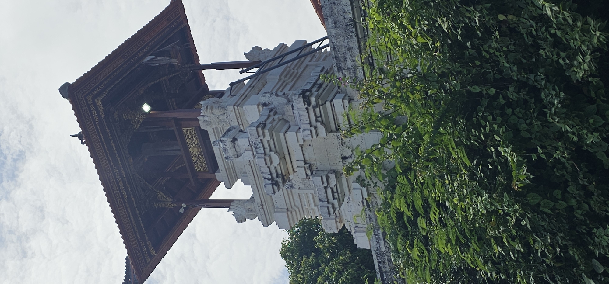 The image shows a temple in Kuta Selatan, Indonesia, partially obscured by lush green foliage. The temple exhibits traditional Balinese architecture with a tiered white stone structure featuring intricate carvings of mythical creatures or deities. Adjacent to it is a dark wooden pavilion with a steeply pitched roof, adorned with detailed gold and red patterns. The sky is visible in the background, with scattered clouds suggesting a partly cloudy day. The environment is a mix of natural and constructed elements, highlighting the blend of culture and nature typical of Balinese landscapes.