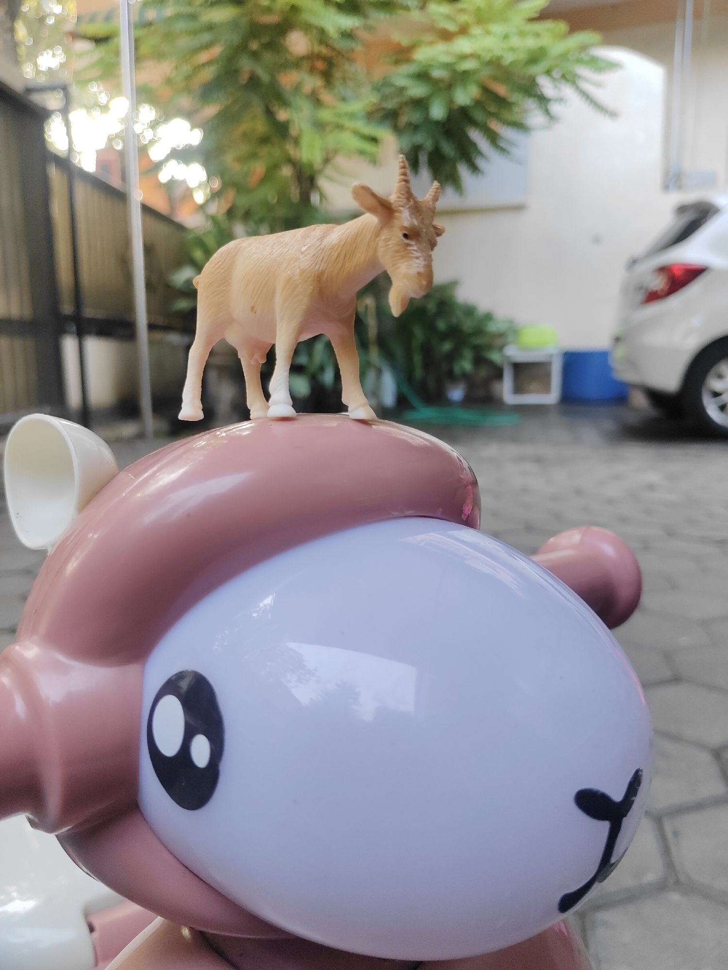 A plastic figurine of a tan goat stands on top of a pink and white toy. The goat is facing to the left and is mid-stride. Its horns are curved backwards. The toy it is standing on is part of a larger toy, possibly a cartoon character with large, black eyes and a smiling mouth. The background is blurred and shows a white car, green trees, a fence, and paved ground. The scene appears to be in an outdoor area, possibly a yard or driveway, in Surabaya, Indonesia.