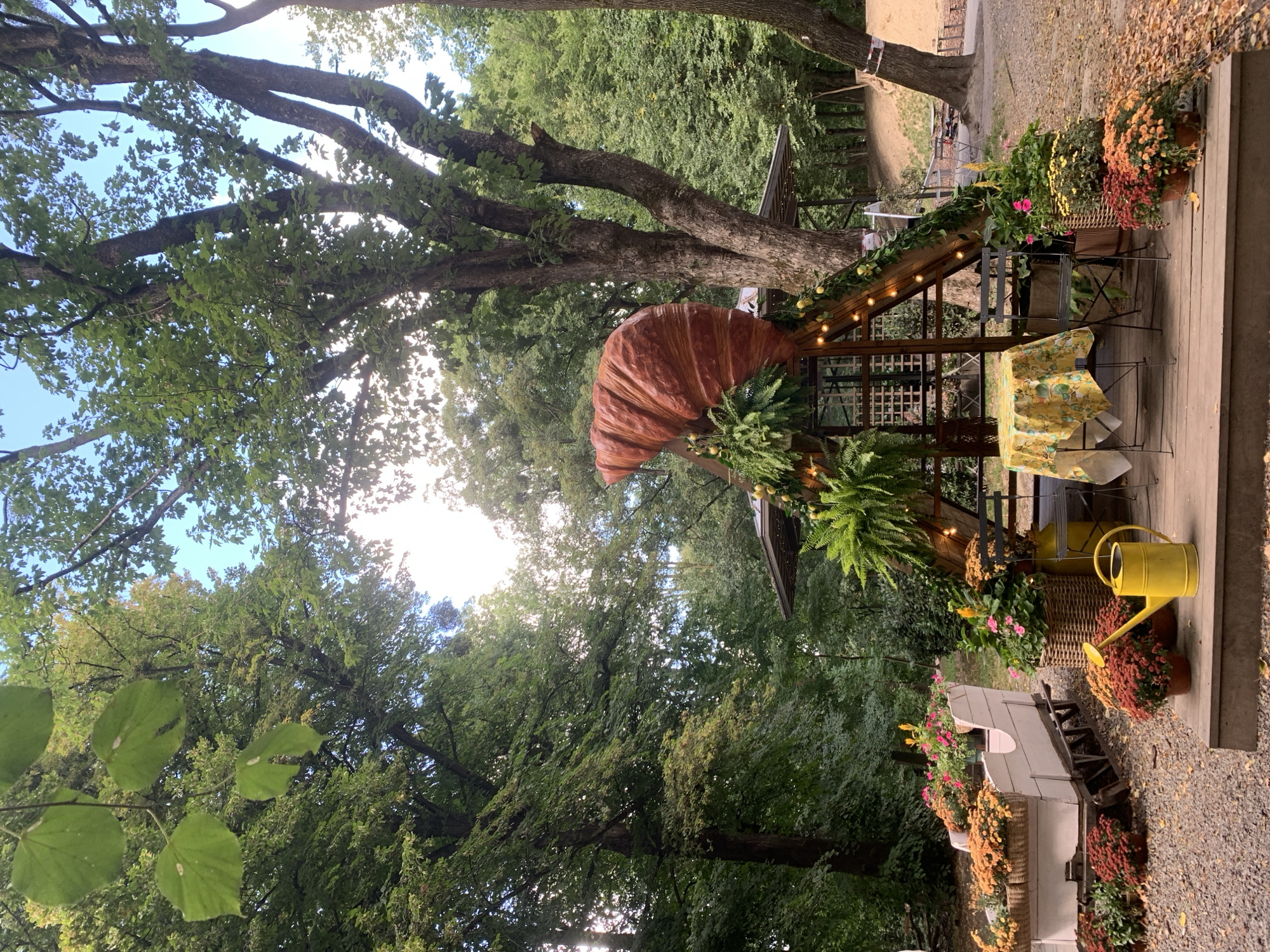 This image depicts an outdoor seating area decorated for autumn, possibly at a cafe or a garden. The scene is set amongst lush green trees, suggesting a park or a wooded area. Prominently featured is a large, decorative croissant sculpture placed atop a rustic wooden structure adorned with string lights and greenery. In the foreground, a white sled-like planter filled with orange and yellow mums sits on a gravel path. A yellow watering can and several potted mums are arranged on a wooden deck. A small table with a yellow, lemon-patterned tablecloth and a metal chair are set up on the deck. The overall ambiance is festive and autumnal. The specific location in Lviv, Ukraine, is not identifiable from the image itself.