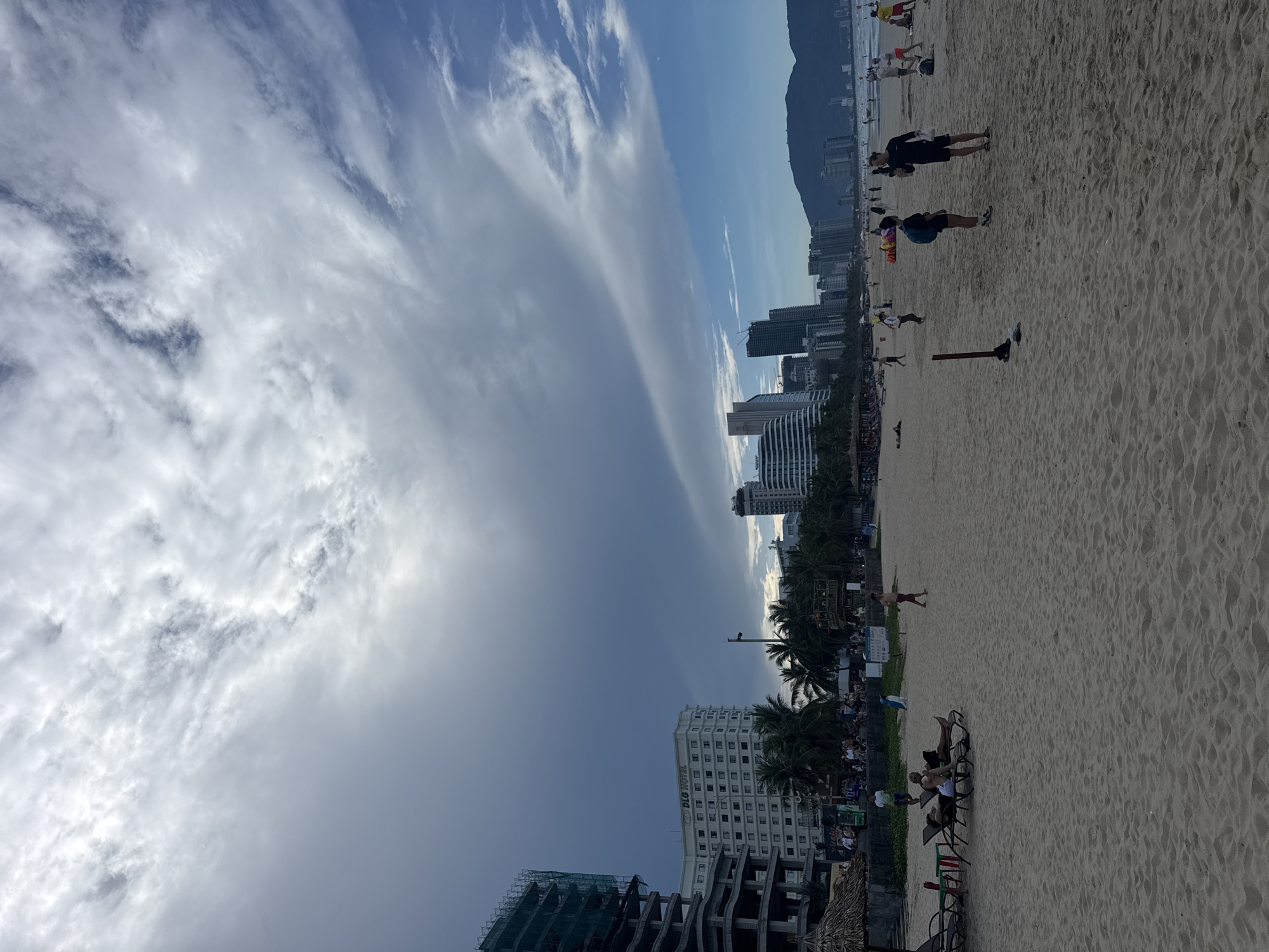 The image captures a beach scene in an unidentified city. The sky features a blend of dark storm clouds and lighter, wispy clouds. In the distance, a mountain range forms a backdrop behind the city skyline. Buildings, including hotels, line the beachfront. The beach is populated with people engaging in various activities such as walking, jogging, and relaxing in beach chairs. Palm trees provide shade along the coastline. An unfinished building is visible near the foreground, adding a contrast to the scenery.