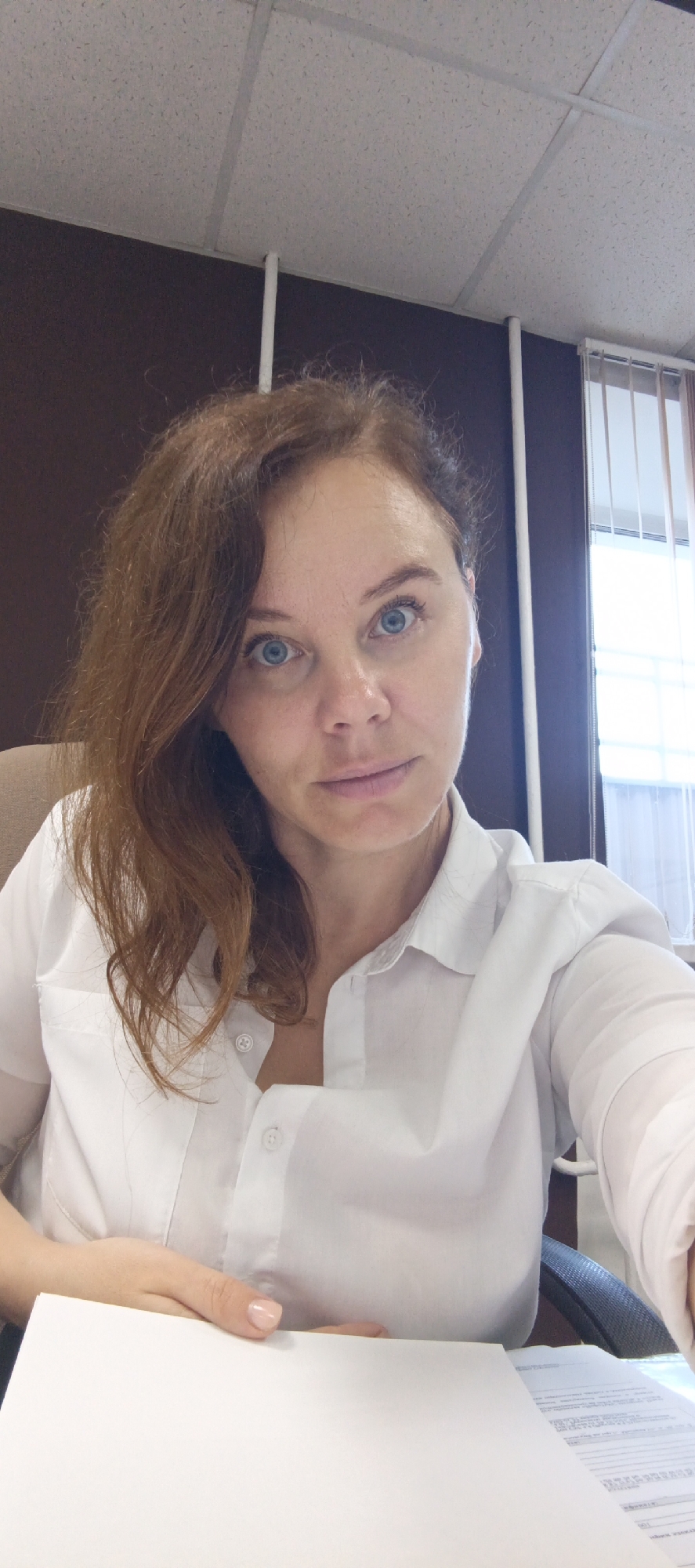 A woman with shoulder-length reddish-brown hair and blue eyes is pictured indoors. She is wearing a white collared shirt. Her left hand rests on a blank white paper on a desk. The background shows a dark wall, a section of a white ceiling with acoustic tiles and metal grid, and a window with vertical blinds. The scene appears to be an office environment. The image is a selfie, taken from a slightly upward angle.