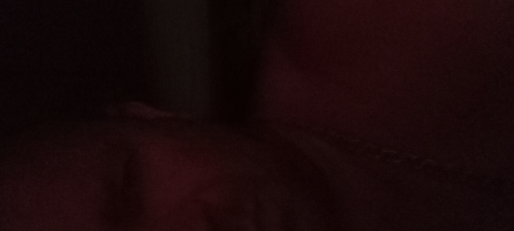 The image is a dimly lit, close-up shot of a person's face, appearing to be taken indoors. The lighting is predominantly red, casting a shadow across the subject's features, obscuring fine details. A silver necklace is visible around the person's neck. The background is dark and indistinct. There is no discernible information that would allow for the identification of the city of Kryvyi Rih, Ukraine, or any specific location within it.