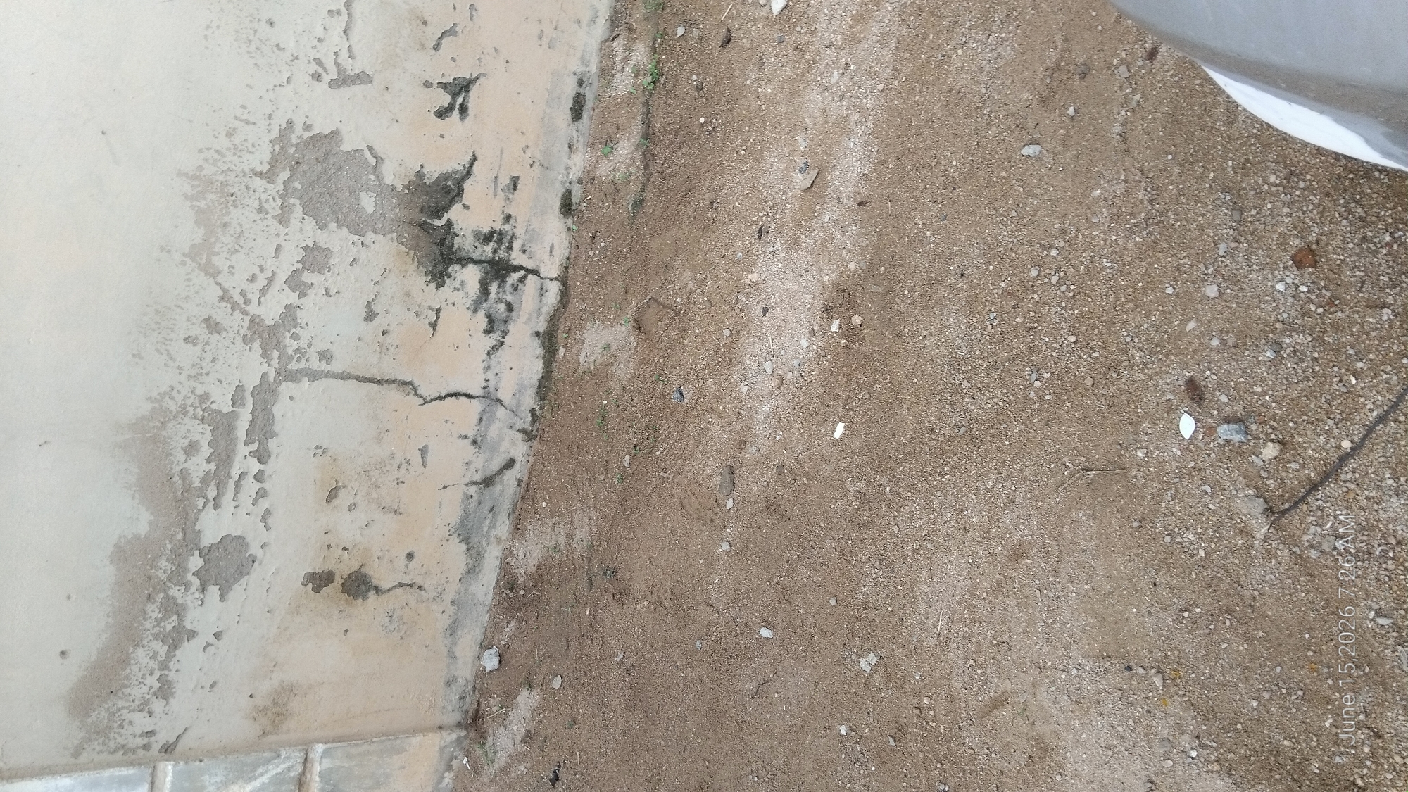 The image depicts an exterior view of a weathered, light-colored wall adjacent to a patch of dry, sandy ground littered with small stones and debris. The wall shows signs of peeling paint and discoloration, with dark streaks and patches that could indicate water damage or mold. A prominent crack runs vertically down the wall in the lower central portion. The ground appears uneven and dry, with a few small green sprigs of vegetation emerging near the wall. The bottom left corner of the image contains a timestamp indicating "June 15 2026 7:26 AM". The image does not provide specific indicators of the exact location within Howayi, Nigeria, beyond being an outdoor setting.