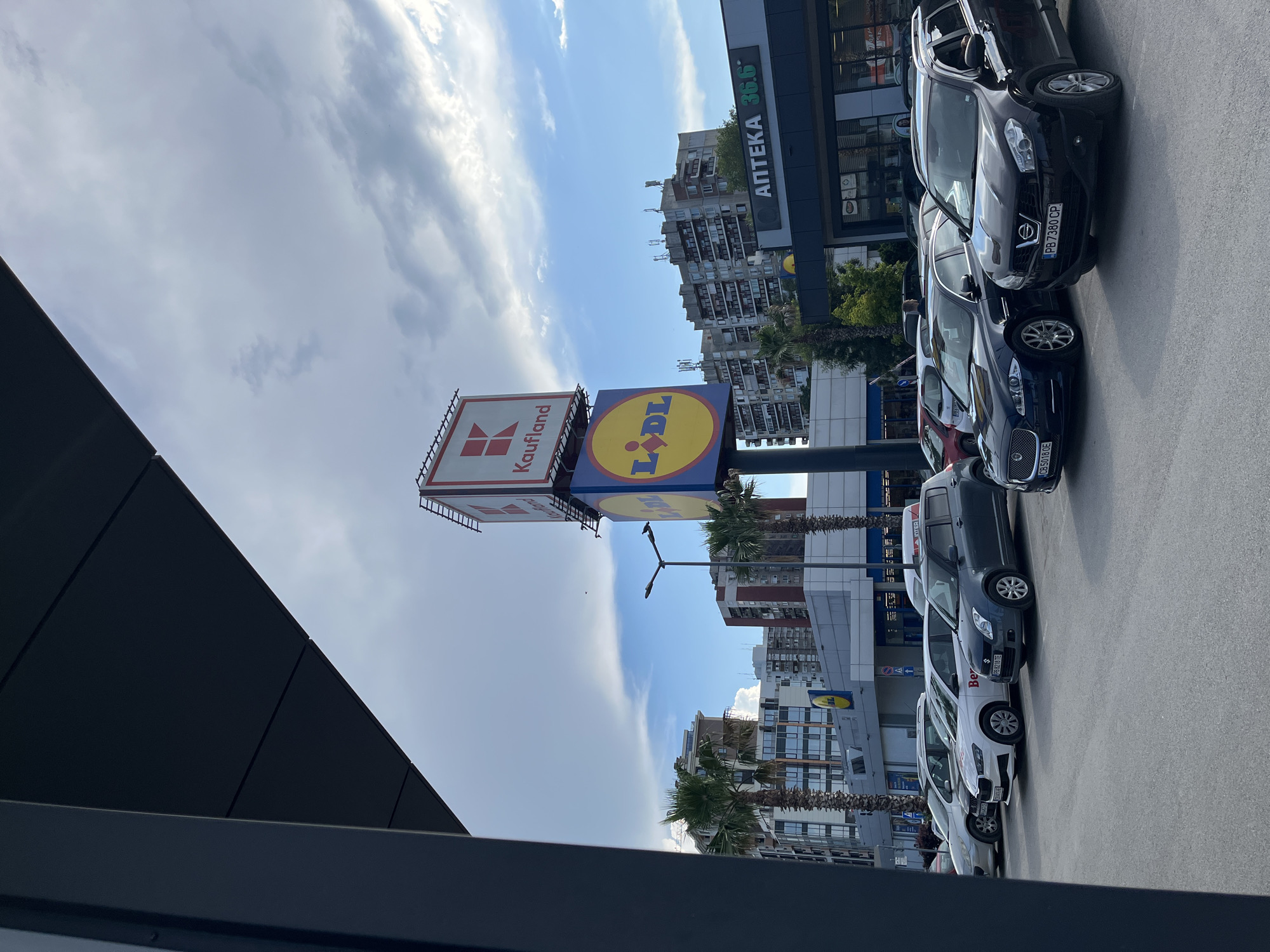 The image shows an outdoor scene with a parking lot in front of retail stores. Several cars are parked in a row. In the background, there are multi-story buildings and palm trees, suggesting a temperate or warm climate. Above the buildings, a cloudy sky with patches of blue is visible.
Prominently displayed are two large signs for retail stores: one for "Kaufland" and another for "LIDL". A third sign indicates "АПТЕКА 36.6", which translates to "Pharmacy 36.6". License plates on some of the cars are visible, with text such as "CB 5018 0E" and "PB 7380 CP". The overall setting appears to be a commercial area or shopping center. The lighting suggests it is daytime, with a mix of sunlight and clouds. The image is oriented vertically, as if taken from a phone held in portrait mode.