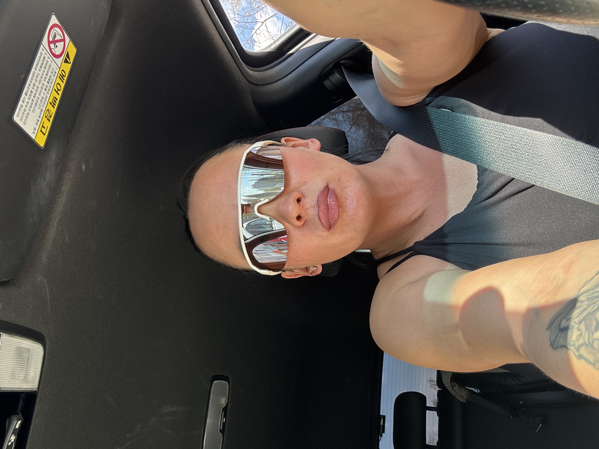 The photograph appears to be a selfie taken inside a car. A woman with short dark hair and wearing sunglasses and a black top is visible. She is wearing a seatbelt. The car's interior is primarily black. A warning label in Korean is visible on the sun visor. A part of a tree is seen through the windshield. The city cannot be seen clearly.