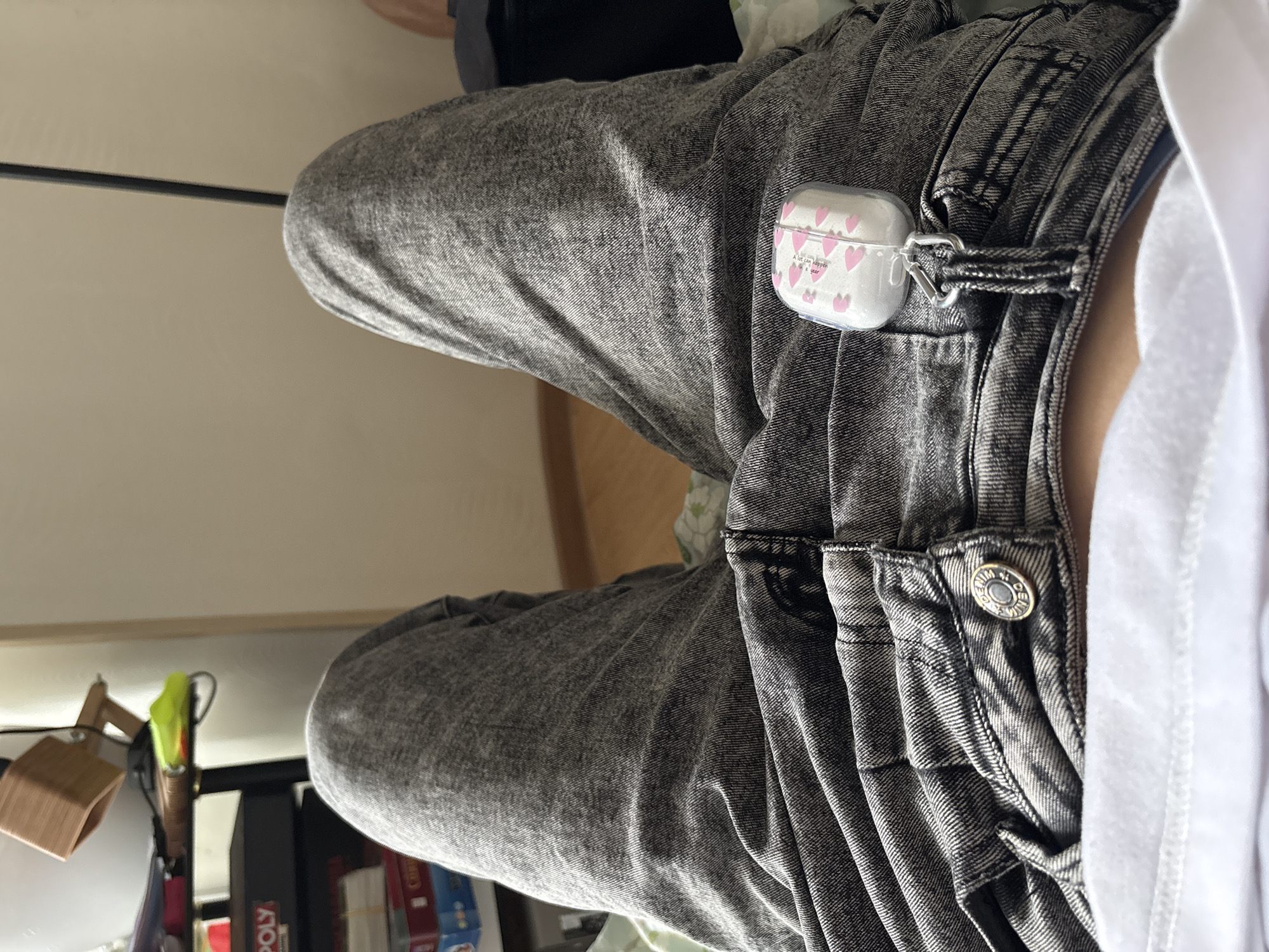 A person is seated, wearing acid-wash jeans. An AirPods case with a pink heart design is clipped to the belt loop. The location is not determined.