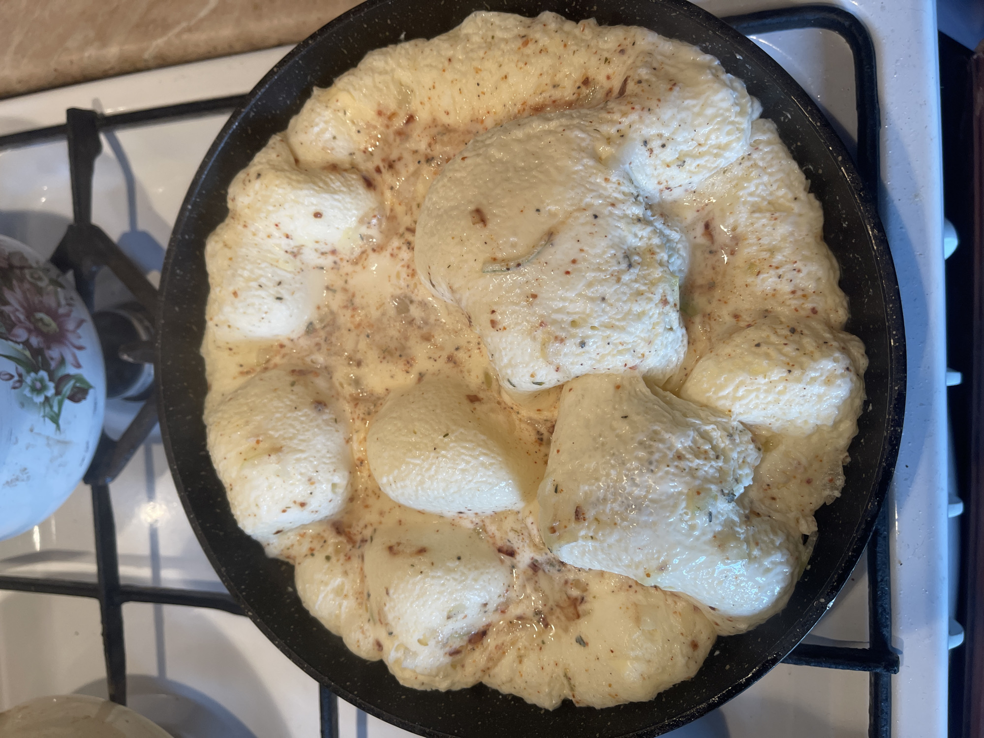 A top-down, close-up shot captures a black frying pan on a white gas stove. Inside the pan, a foamy, creamy substance, likely scrambled eggs or a similar dish, is being cooked. The mixture is a pale yellow, with visible specks of herbs and spices, including what appear to be red pepper flakes and dried herbs. Several lighter-colored, uneven lumps are dispersed throughout the mixture, suggesting they might be portions of cheese or some other solid ingredient that is melting or being incorporated. The overall texture of the dish appears soft and somewhat liquid. The pan itself has a textured, non-stick surface. In the background, on the left, a white pitcher with a floral pattern is partially visible, adding a domestic touch to the scene. The stove's grates and burners are also visible, indicating the dish is actively being cooked. The lighting suggests it is daytime, and the setting appears to be a home kitchen. There is no visible text in the image.