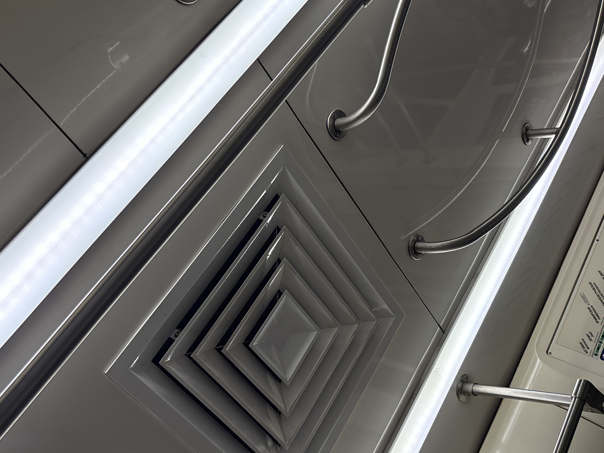 The image displays the clean, modern interior ceiling of a metro car, shot from a low, upward-looking angle, with the composition tilted diagonally. The central feature is a multi-layered, stepped square air vent, providing a strong sense of depth and geometric pattern.
Bright, elongated fluorescent light fixtures run parallel to the length of the car, illuminating the grey and white ceiling panels. Several polished metal handrails, curved for passenger grip, are prominently featured, reflecting the bright overhead lights. The surfaces of the panels and fixtures appear largely clean and well-maintained. A faint, light-colored streak or residue is visible on one of the ceiling panels.
On the right side of the frame, a portion of a route diagram or map is visible on the wall. This map features Cyrillic text, with station names such as "ПРИМОРСКАЯ" (Primorskaya), "ЗЕНИТ" (Zenit), and "БЕГОВАЯ" (Begovaya) clearly decipherable. These names correspond to stations on the Saint Petersburg Metro system, confirming the location as Saint Petersburg, Russia.
No people are visible in the image, and no specific activity is taking place beyond the implied travel within a public transport system. The bright artificial lighting suggests an indoor setting, typical of a metro environment, without indicating a specific time of day.