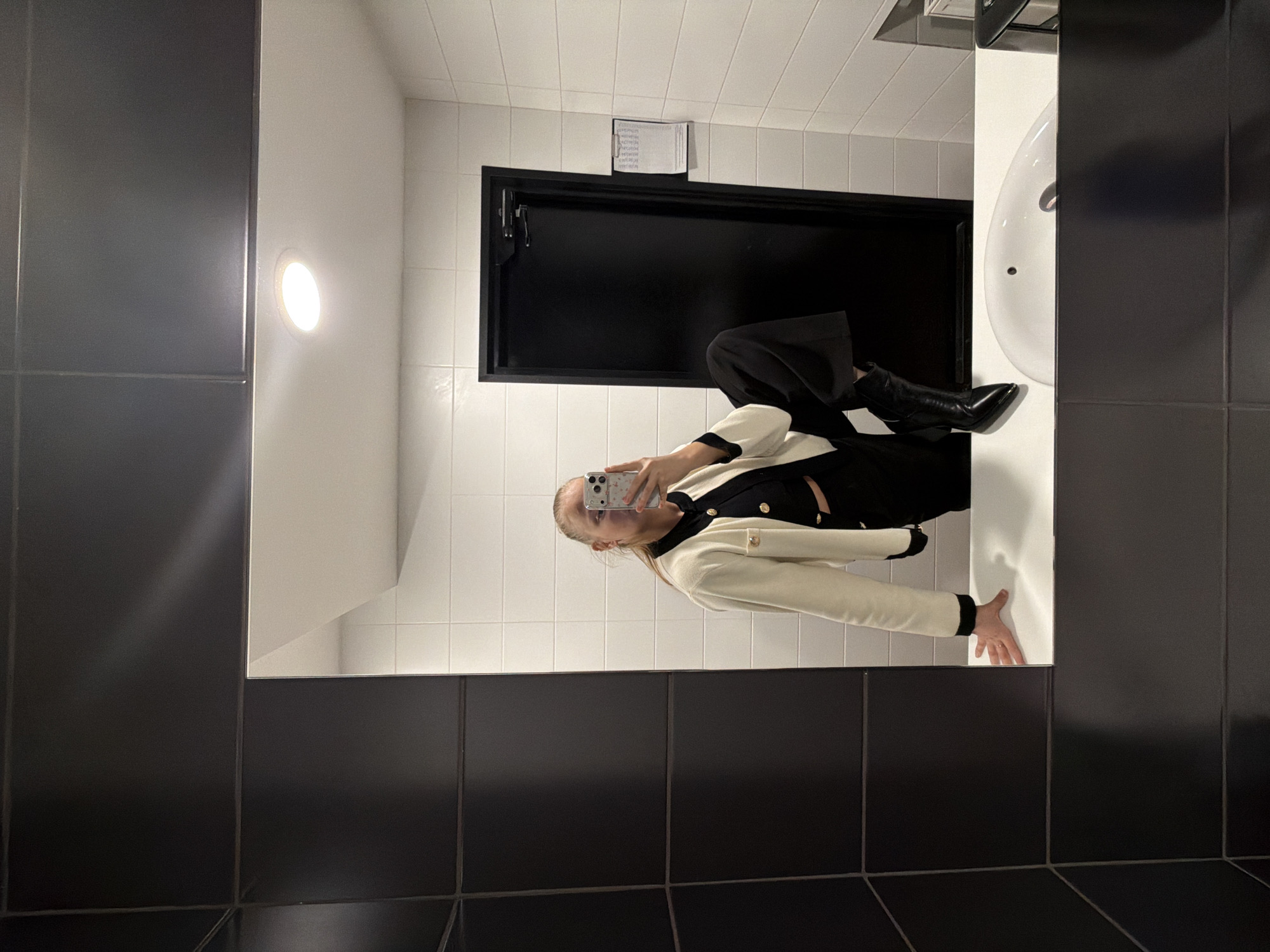 A young woman is taking a selfie in a bathroom mirror. The bathroom has black and white tiled walls. The woman is wearing black pants, black boots, and a white jacket with black trim. She is holding a cell phone in her left hand. There is a sink and a black door in the background. The photograph was not taken in Izhevsk, Russia.