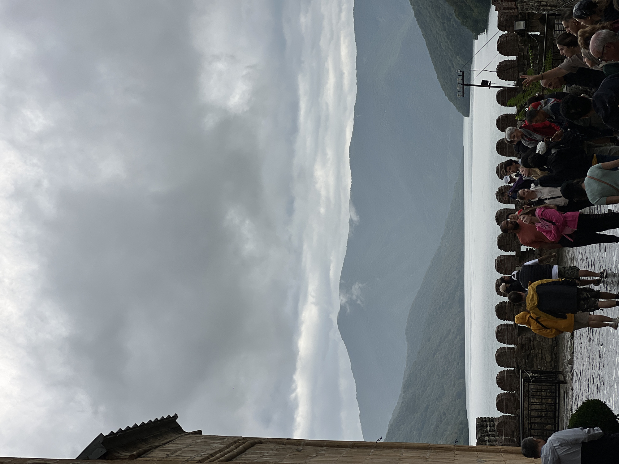 A group of tourists stands on a stone-paved courtyard of a historical complex, possibly Ananuri Fortress, in Pkhundavi, Georgia. The tourists, dressed in casual outdoor wear, are looking out at a vast body of water, likely the Zhinvali Reservoir, surrounded by lush green mountains. The sky is overcast with heavy, gray clouds, creating a dramatic and somewhat muted atmosphere. The architectural style of the courtyard, with its stone walls and crenellated battlements, suggests an ancient defensive structure. Some individuals are pointing or gesturing, possibly engaging with a tour guide or observing the scenery. The overall scene captures a moment of exploration and appreciation of the natural and historical landscape of the region.
