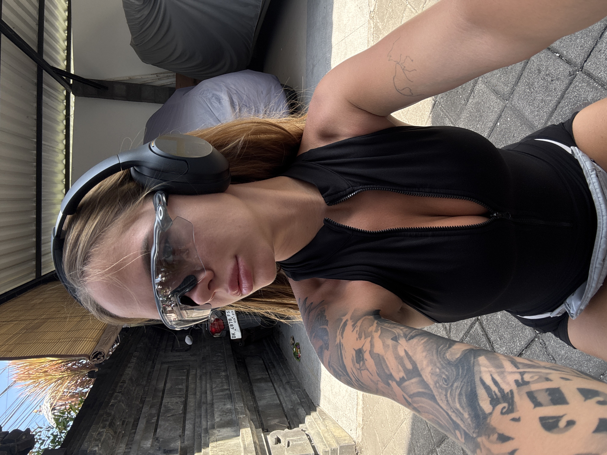 The image shows a woman wearing safety glasses and headphones. She is dressed in a black top and shorts, and has tattoos on both arms. She is standing on a paved surface. Behind her, a vehicle license plate reads "DK 4878 FS". The background suggests an outdoor setting with architecture that may be indicative of North Kuta, Indonesia. The image is shot from a first-person, selfie perspective.