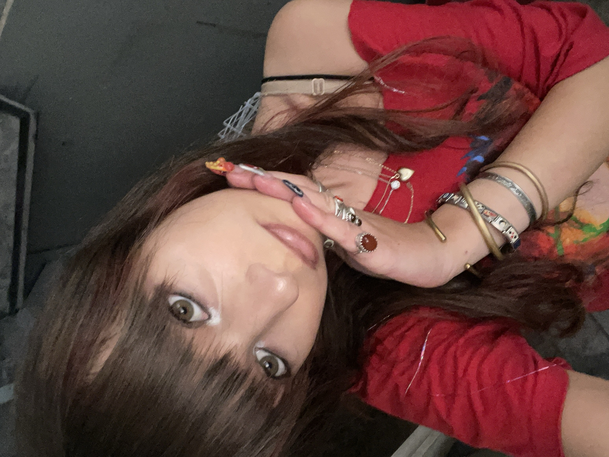 In this medium shot photograph, a young woman with long, dark brown hair and bangs is positioned in the foreground, her face angled towards the camera. She is wearing a red off-the-shoulder top. Her left hand is raised to her face, with her fingers gently touching her cheek. Her fingernails are long and decorated with colorful designs, including what appears to be a miniature figurine on one nail. She is adorned with multiple rings on her fingers and several bracelets on her wrist, including bangles and a charm bracelet. Around her neck, she wears two delicate necklaces, one with a heart-shaped pendant. The background is dark and out of focus, suggesting an indoor environment. The scene is devoid of any recognizable landmarks that could pinpoint a specific location in Raanana, Israel.