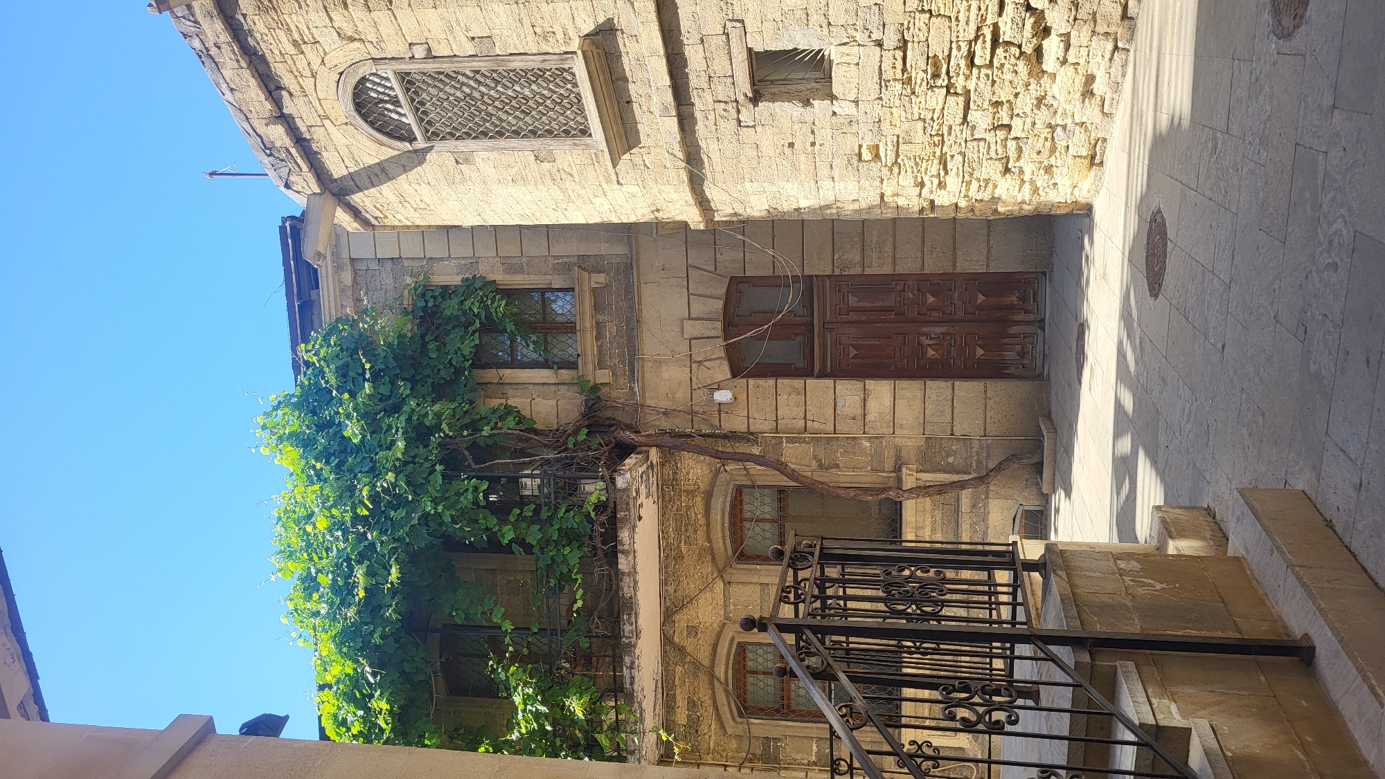 The image depicts a sunlit courtyard in Baku, Azerbaijan, characterized by old stone architecture. A building with a yellow stone facade dominates the scene, featuring multiple windows with decorative metal grilles and a large, ornate wooden double door. A thick vine, laden with vibrant green leaves, climbs the facade of the building, creating a natural canopy over a balcony and extending down the wall. Adjacent to this building, another structure, also made of light-colored stone, is visible on the right, with a prominent arched window and a smaller, square window. The courtyard floor is paved with large, square stone tiles, and shadows from the structures and vegetation create a dappled pattern across the ground. A black metal railing and steps are present in the foreground on the left, suggesting an entrance or pathway. The sky above is a clear, bright blue. The overall impression is one of historical charm and a tranquil, sun-drenched atmosphere.